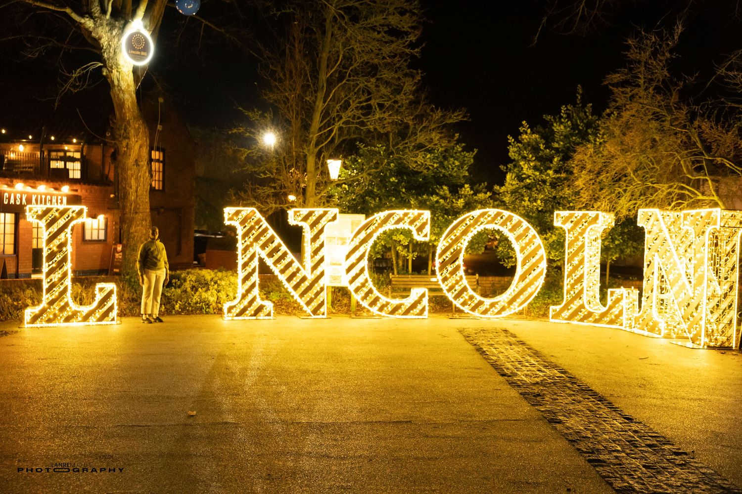 Illuminated LINCOLN sign glowing golden at night in an outdoor plaza with trees and buildings in background.