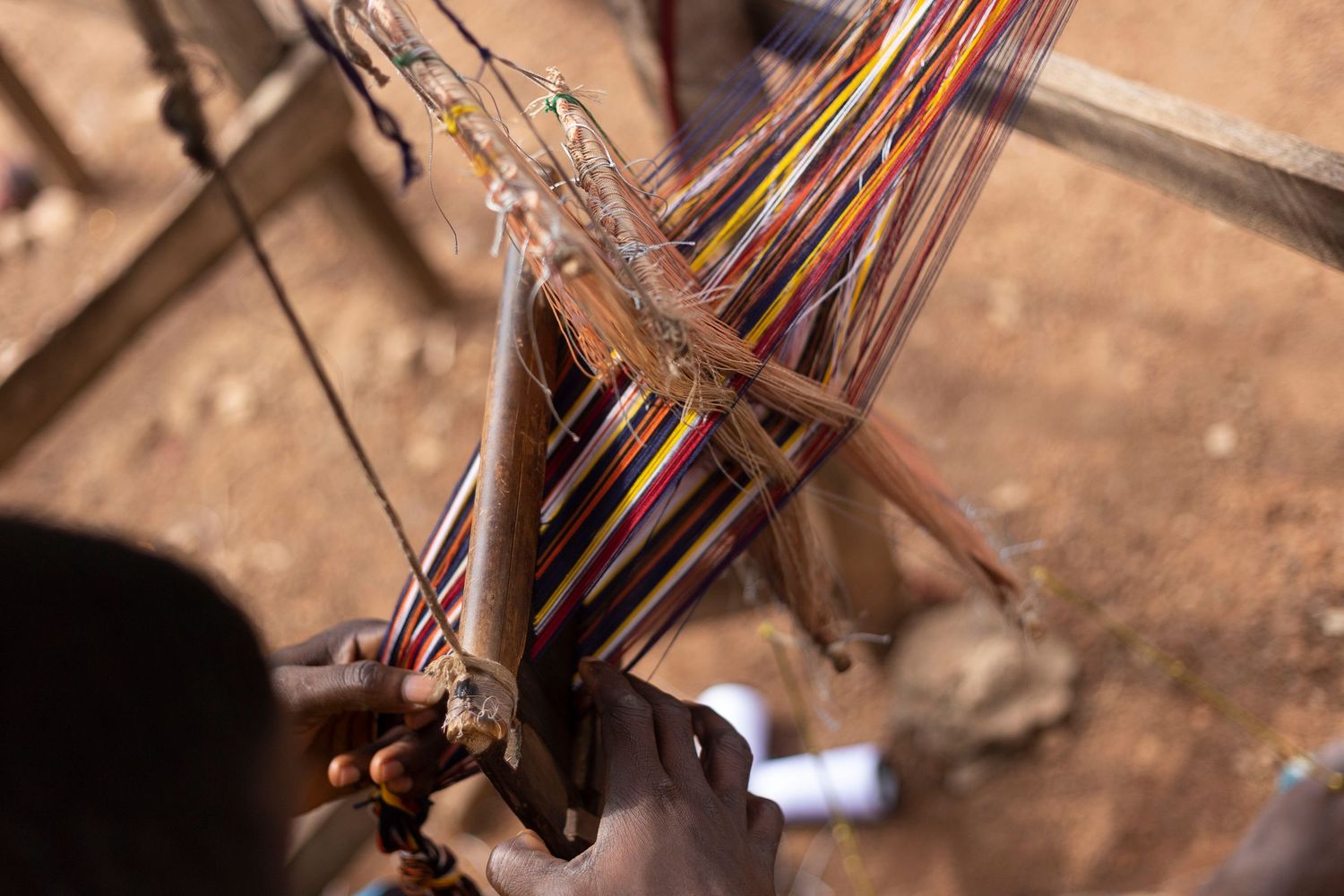 Colorful threads on a traditional hand loom being woven by a craftsman on a dirt ground.