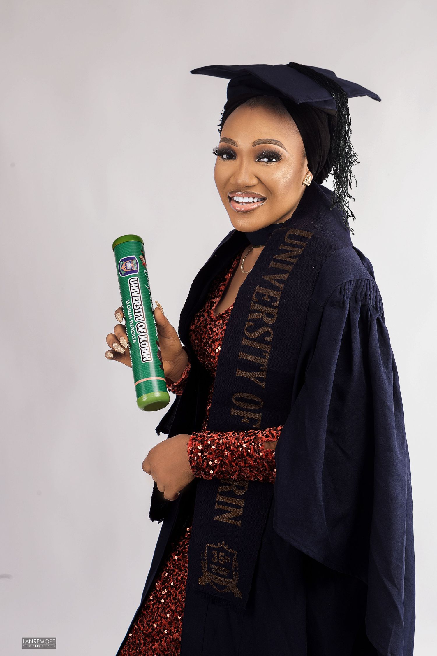 Graduate woman in cap and gown with University of Ilorin sash holding diploma in studio portrait.