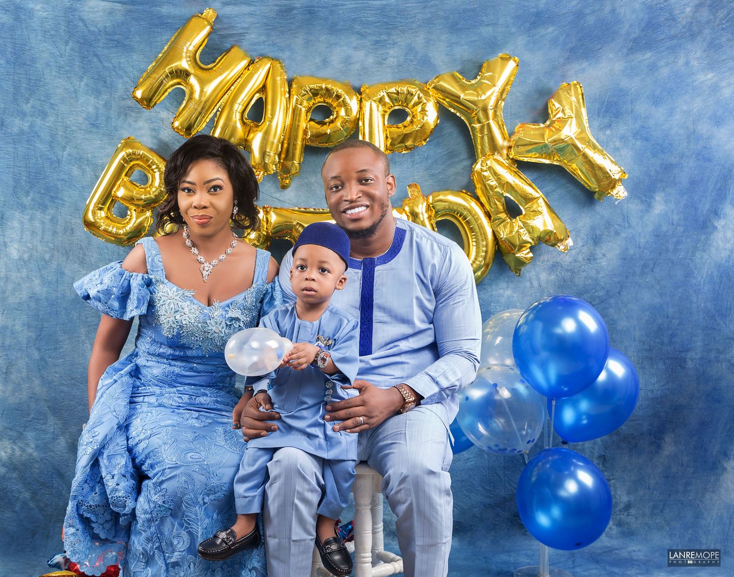 Family celebrating baby's first birthday in blue outfits with Happy Birthday gold balloon letters.