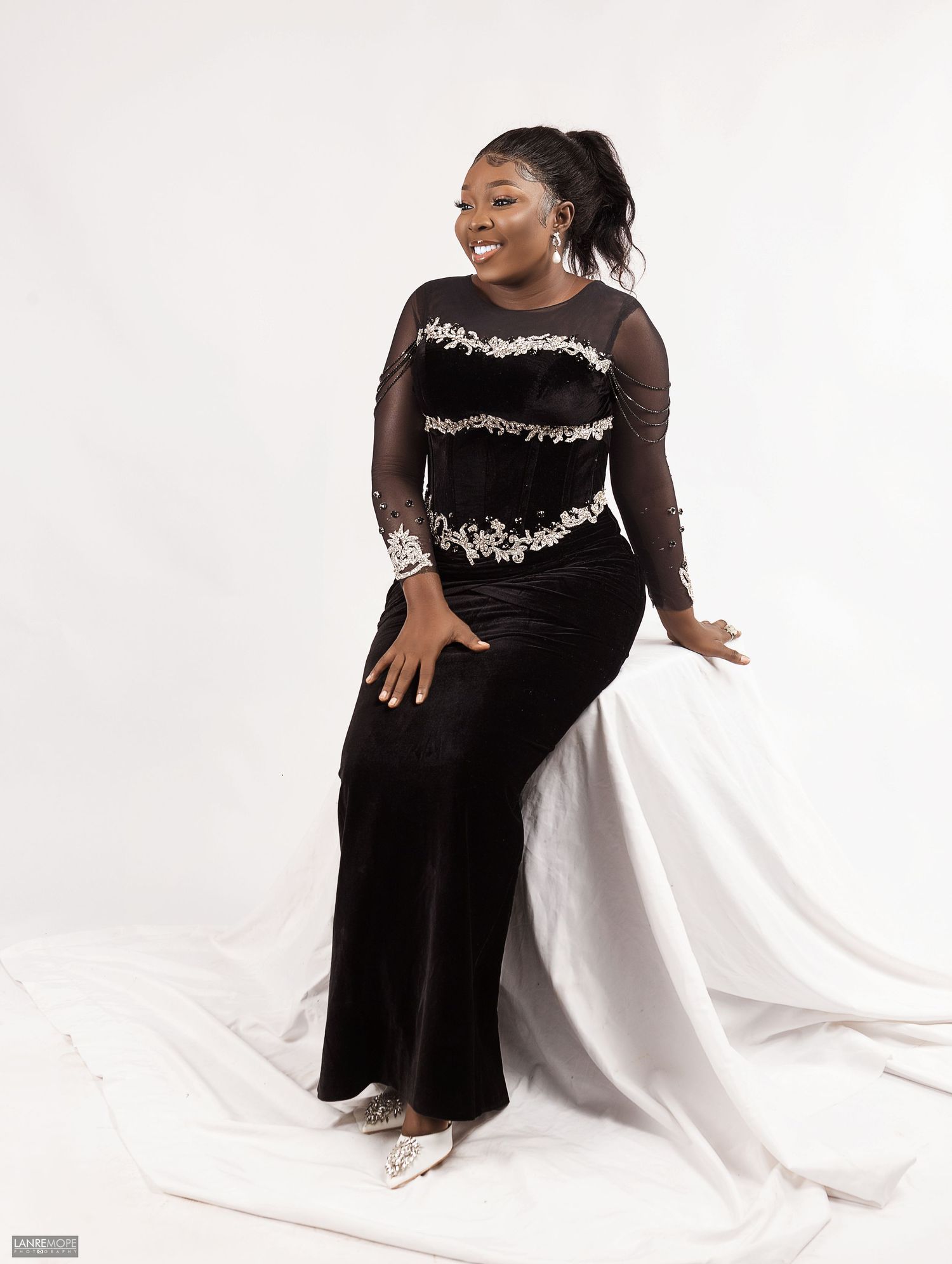 Woman in black velvet embroidered formal gown seated elegantly in professional portrait studio photo.