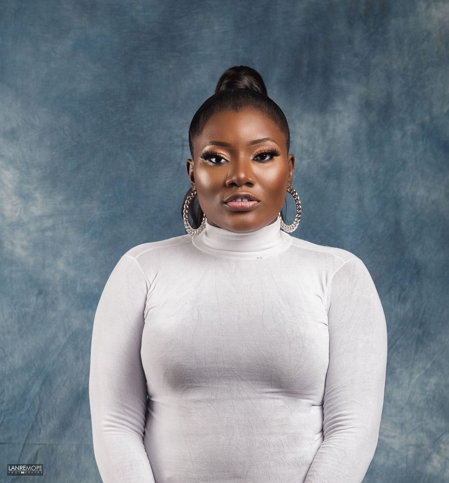 Woman in white turtleneck dress with hoop earrings posing against blue-gray studio backdrop.