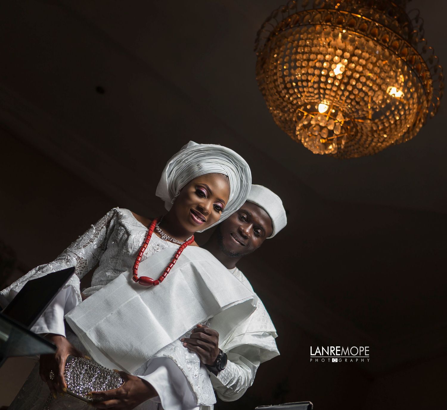 Nigerian couple in white traditional wedding attire posing under golden chandelier.