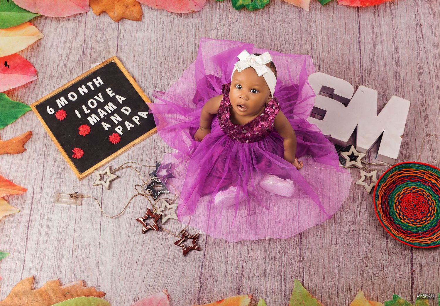 6-month-old baby girl in purple tutu dress lying on wooden floor with autumn leaves and star props.