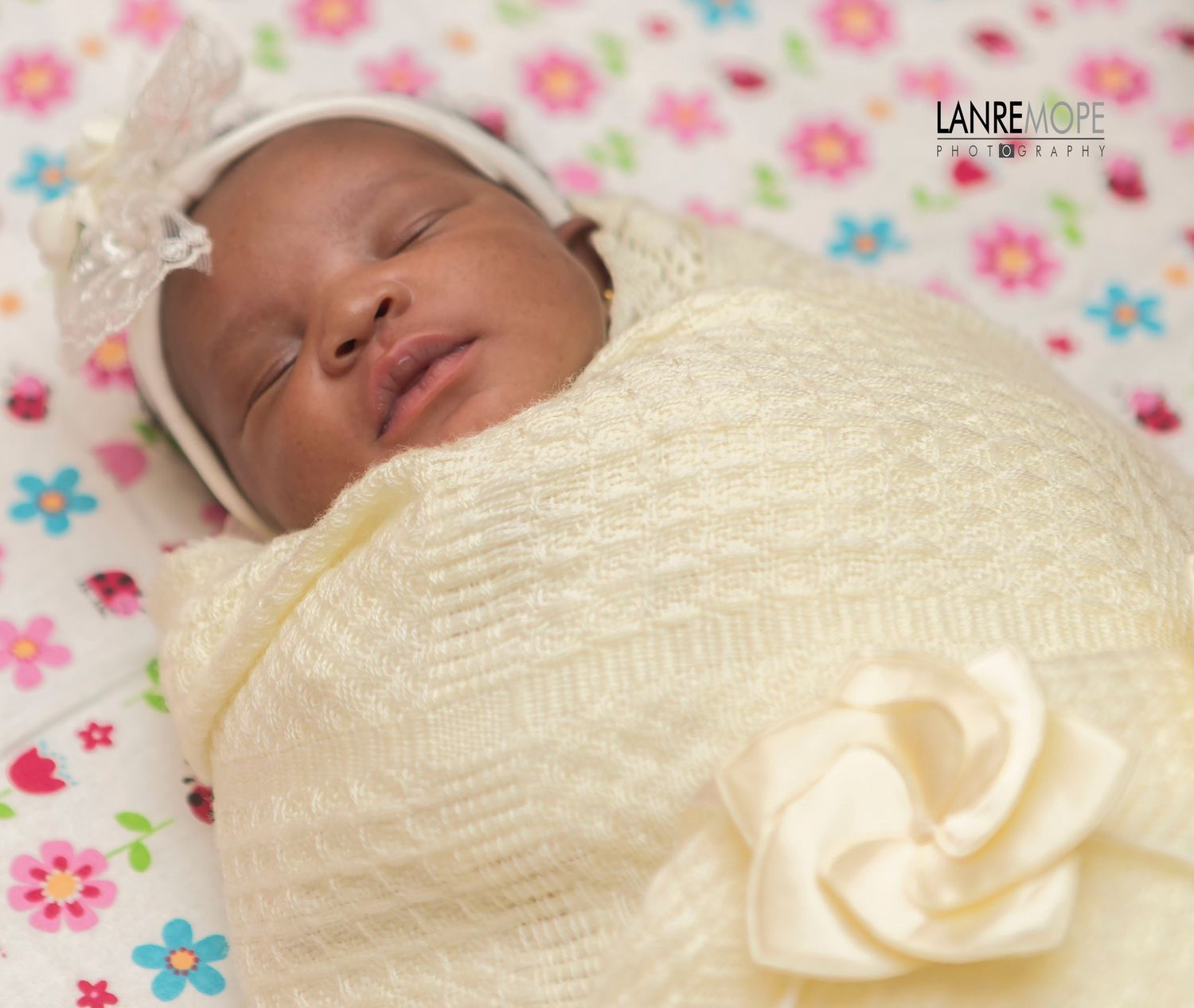 Newborn baby swaddled in cream knit blanket with white bow headband, sleeping peacefully on floral print surface.