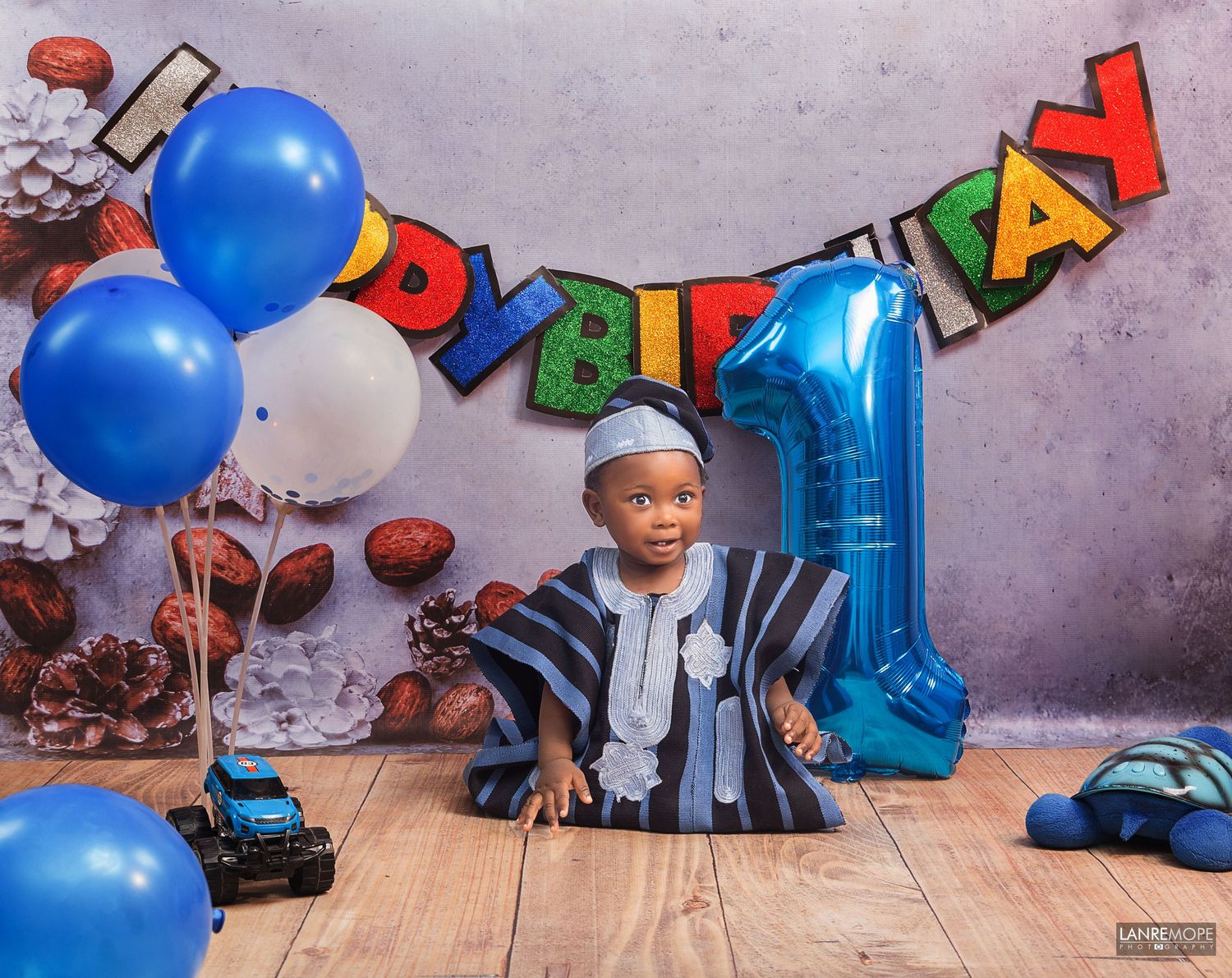 Baby boy in traditional Nigerian attire celebrating first birthday with colorful balloons and number 1.