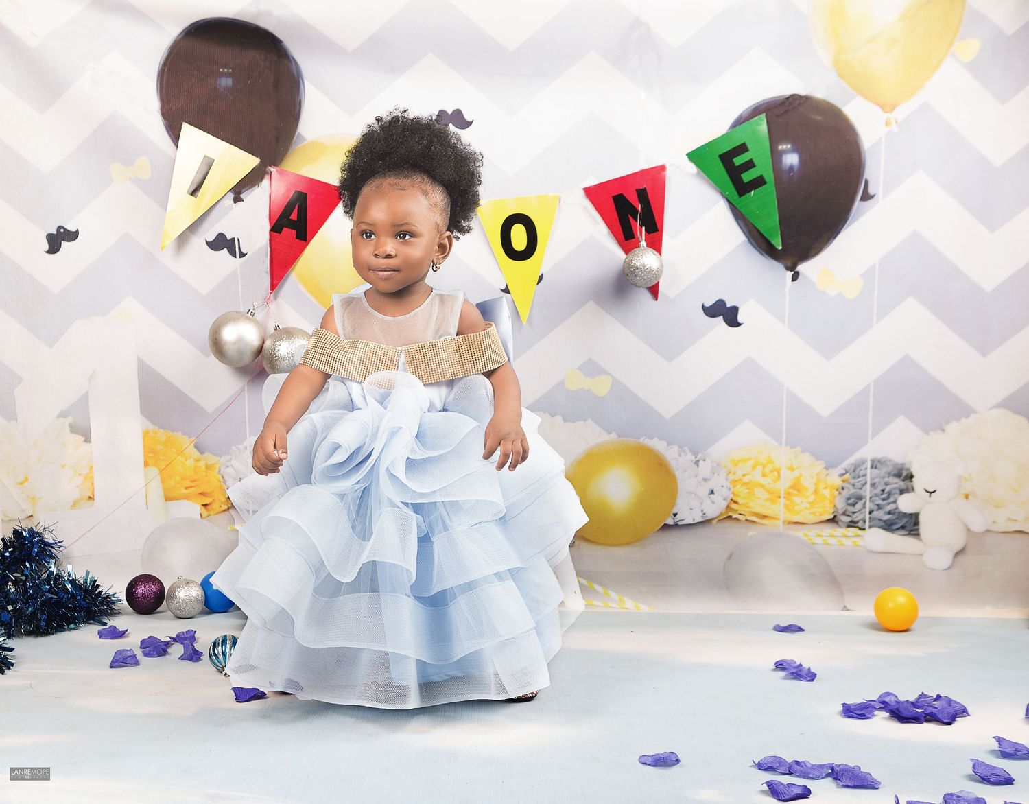 Baby girl in blue ruffled dress standing among balloons and confetti at a first birthday celebration.