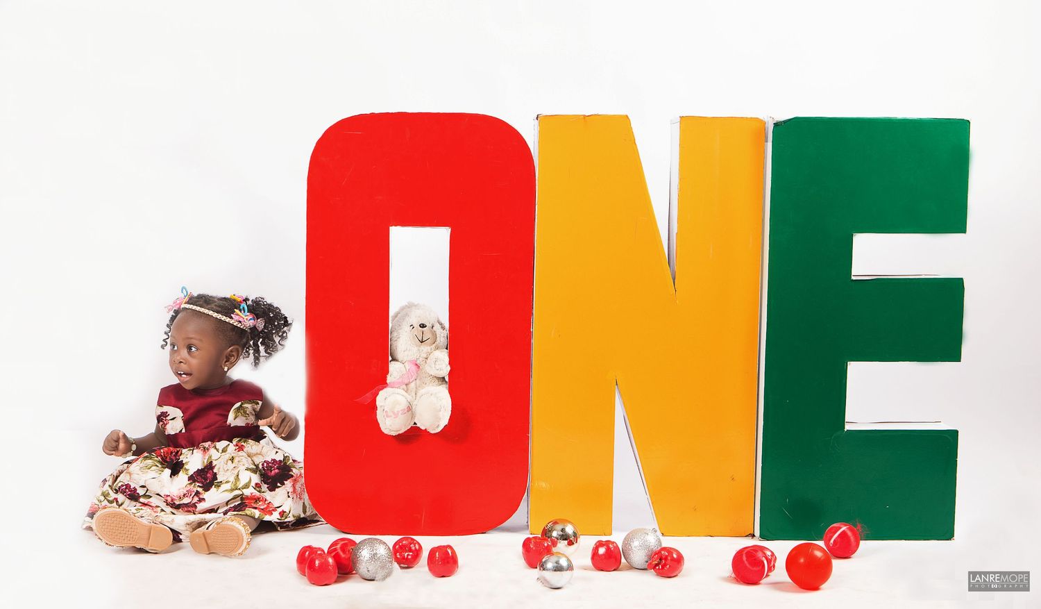 Baby girl in floral dress sitting beside a teddy bear next to colorful ONE birthday letters.