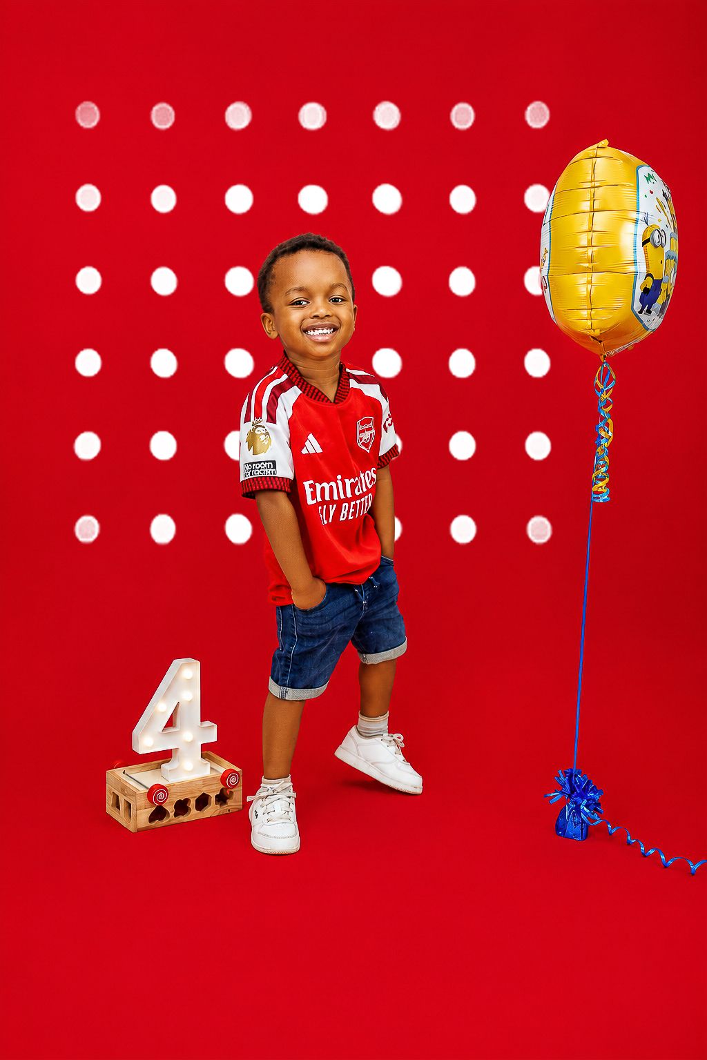 Smiling boy in Arsenal jersey celebrating 4th birthday with gold balloon on red studio background.