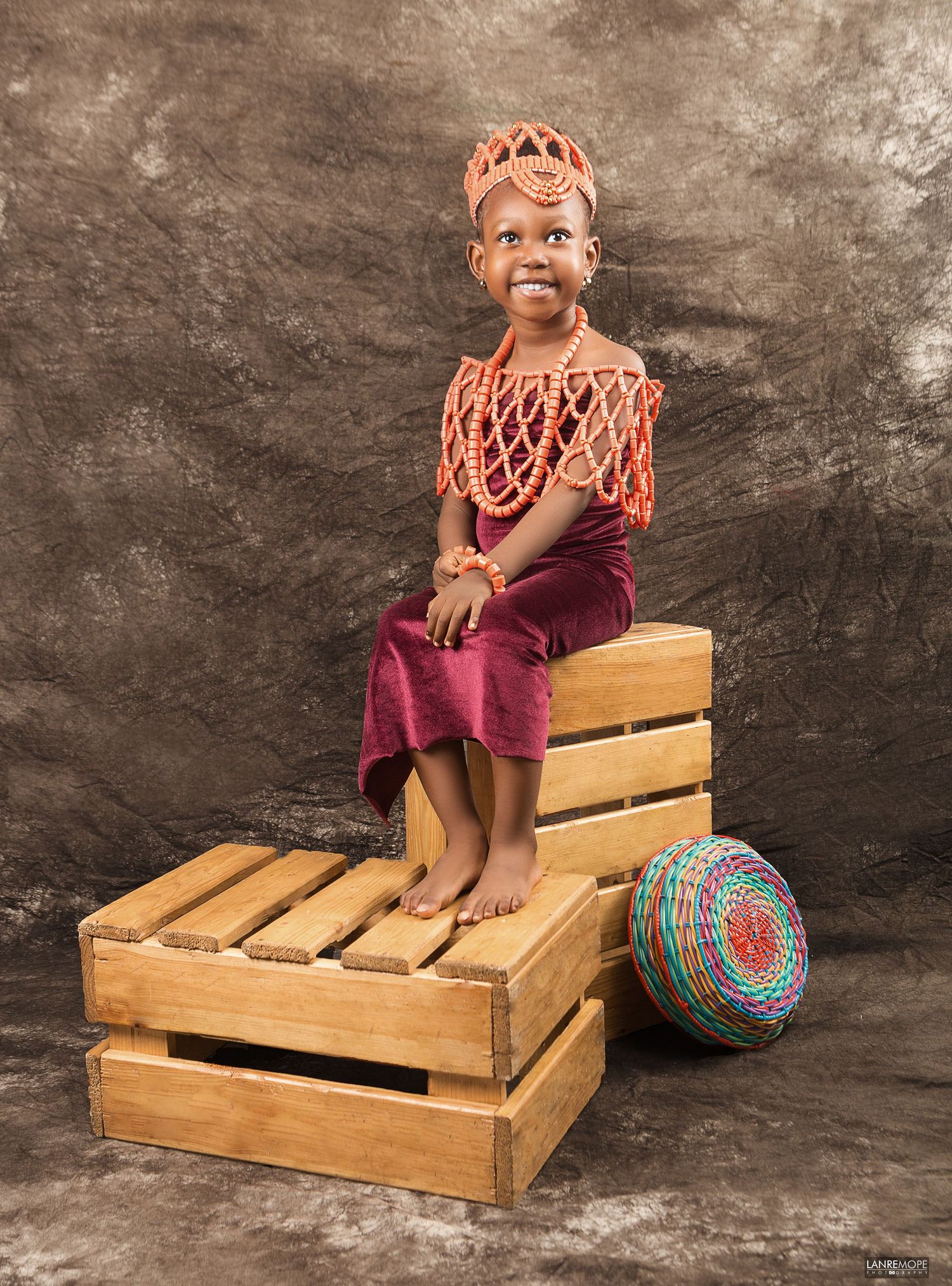 Young Nigerian girl in traditional coral beaded attire seated on wooden crates in a studio portrait.