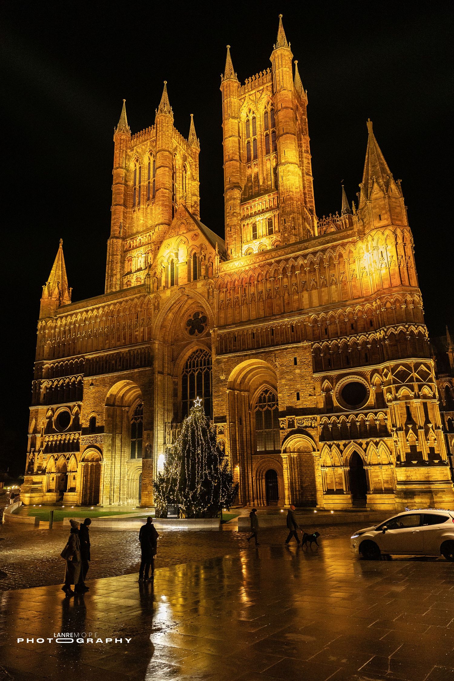 Lincoln Cathedral illuminated at night with a Christmas tree in the foreground reflecting on wet cobblestones.