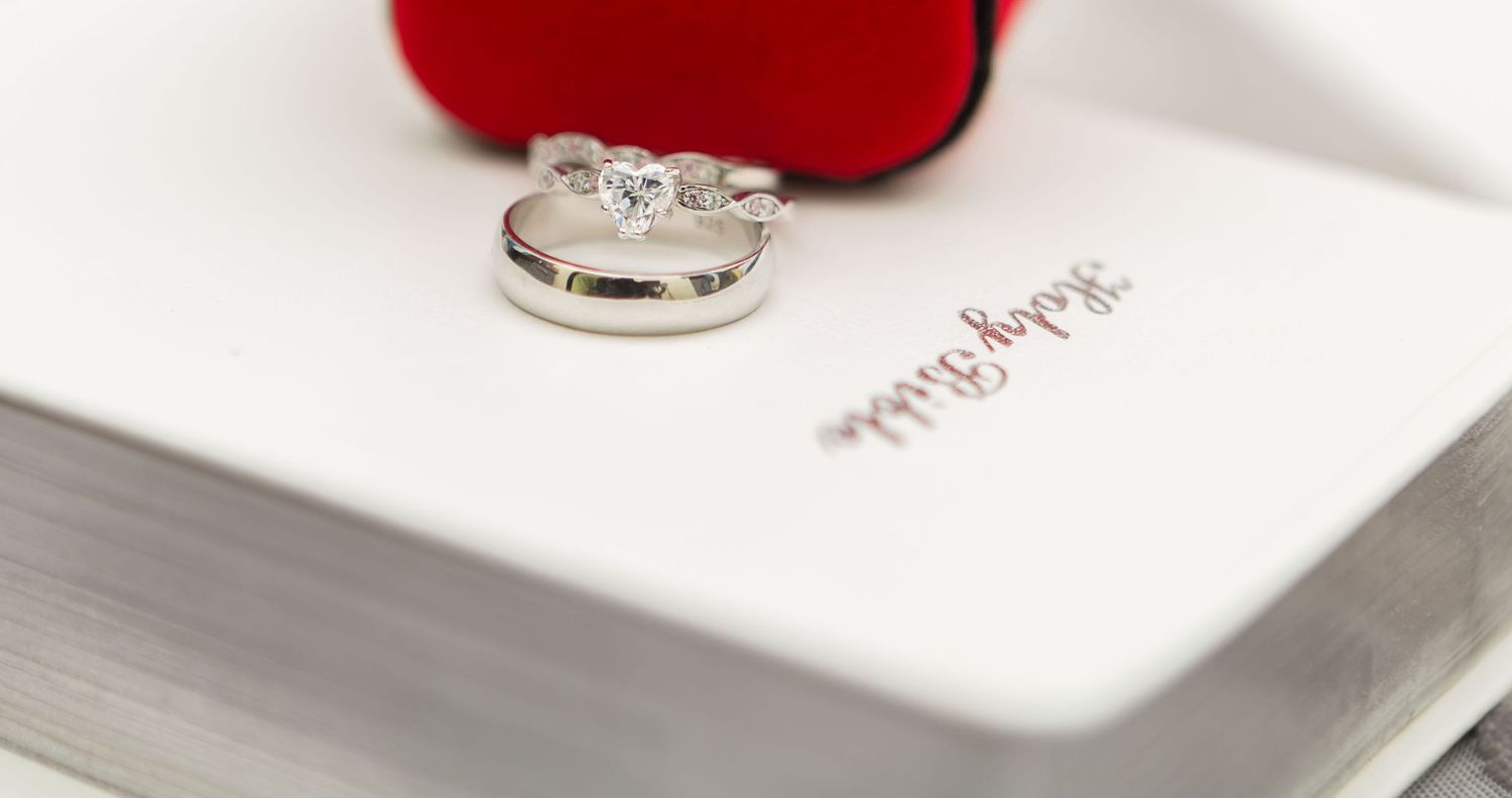 Diamond engagement ring and wedding band resting on a Holy Bible with red ring box.