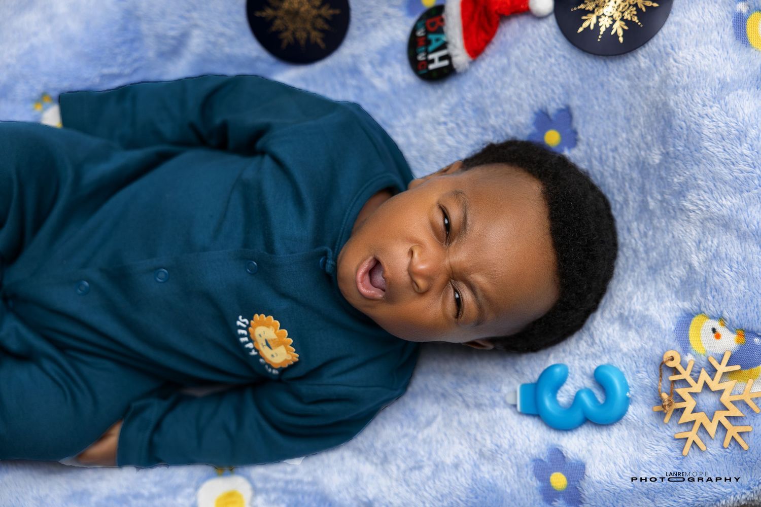 Adorable baby boy in teal outfit lying on a blue blanket with holiday decorations around him.