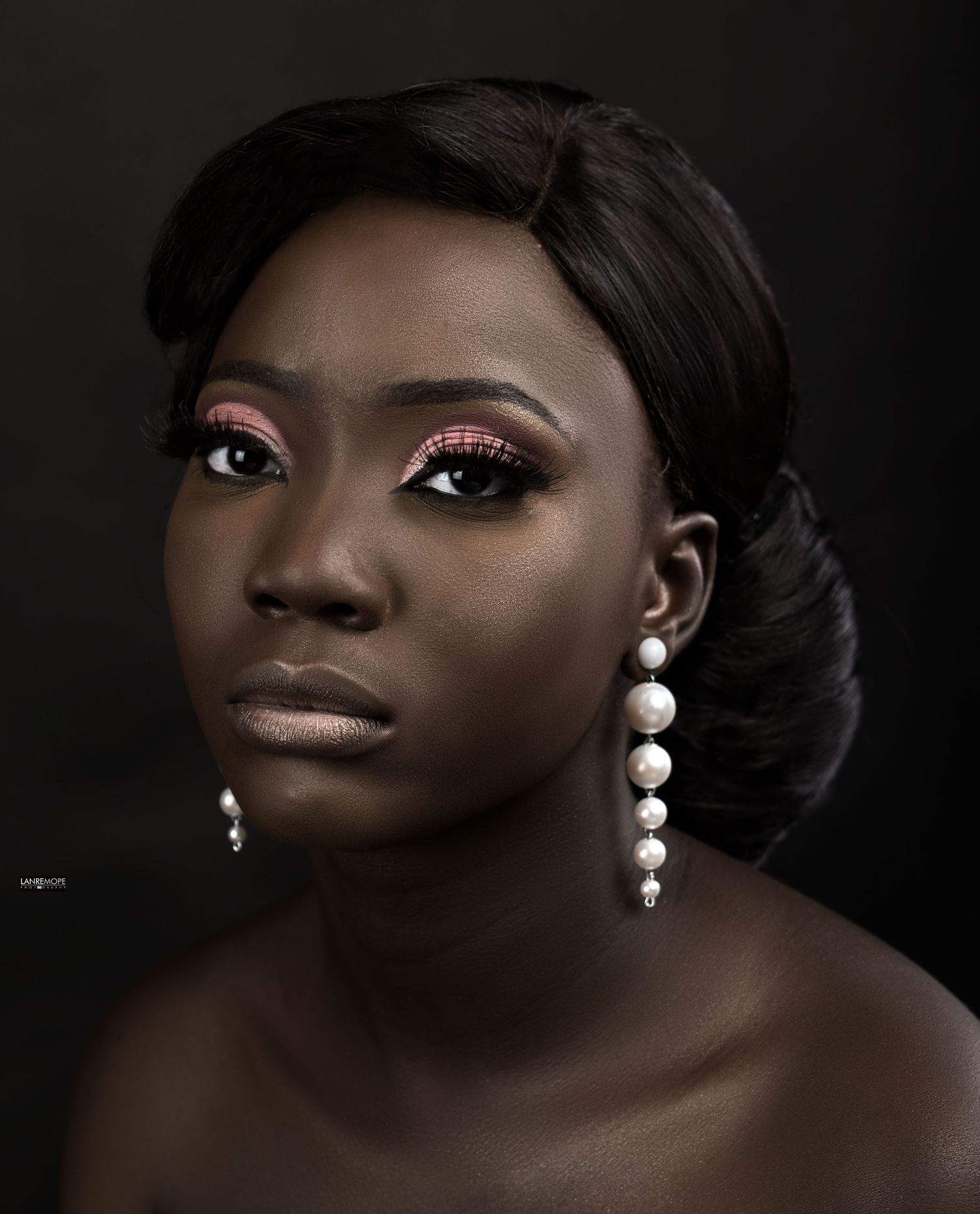 Close-up portrait of woman with dramatic eye makeup and pearl drop earrings on dark background.
