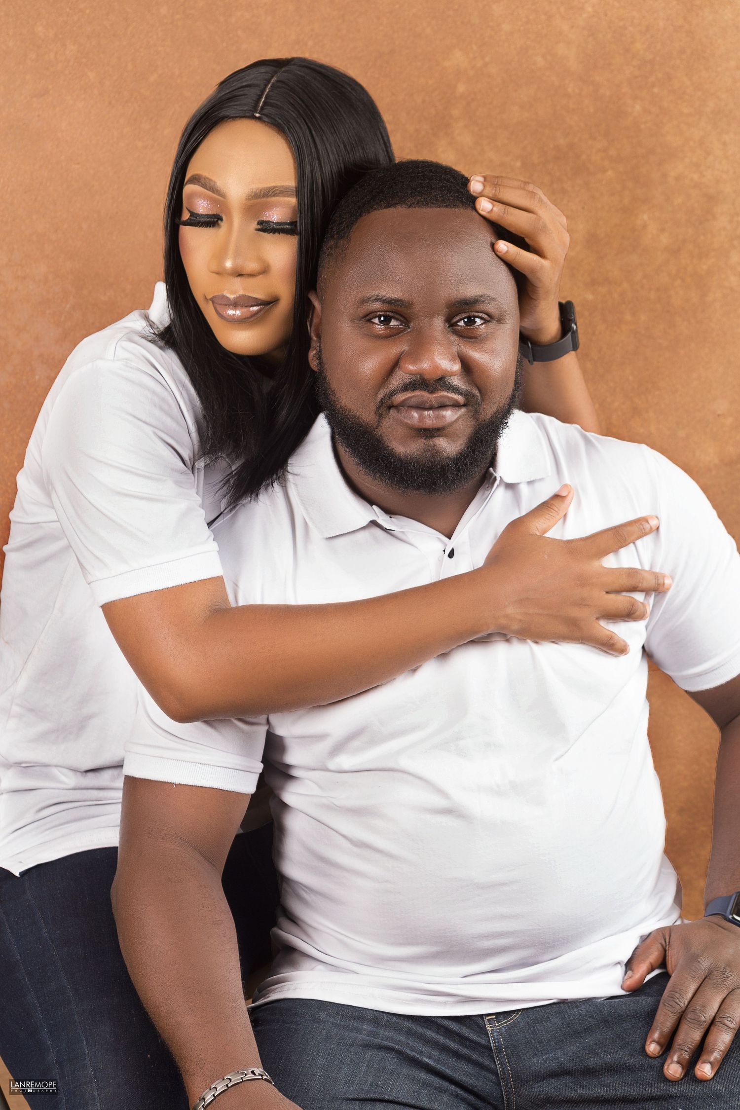 Couple posing in matching white polo shirts, woman embracing man from behind in a studio portrait.