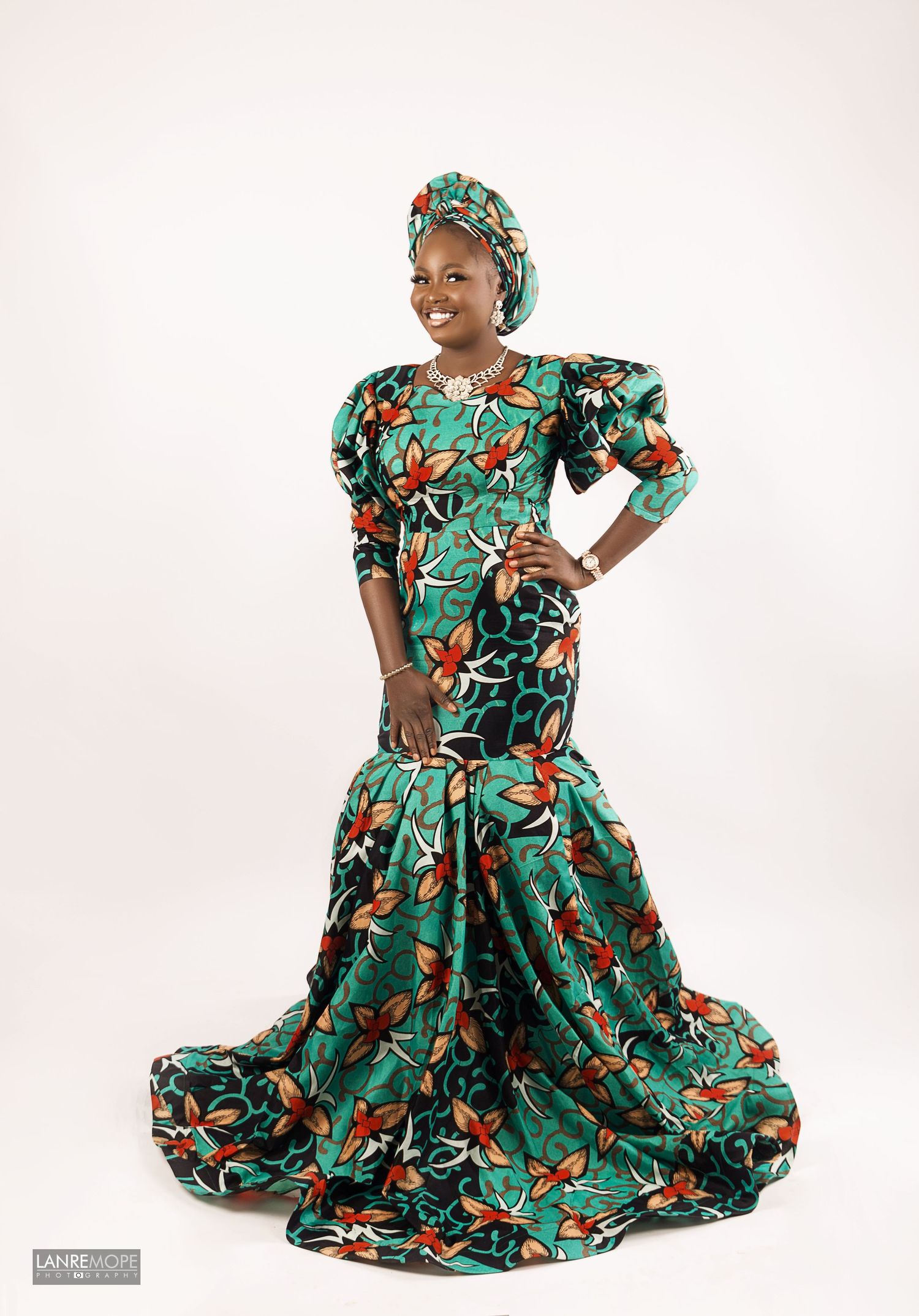 Woman posing elegantly in green floral African ankara mermaid gown with matching head wrap.
