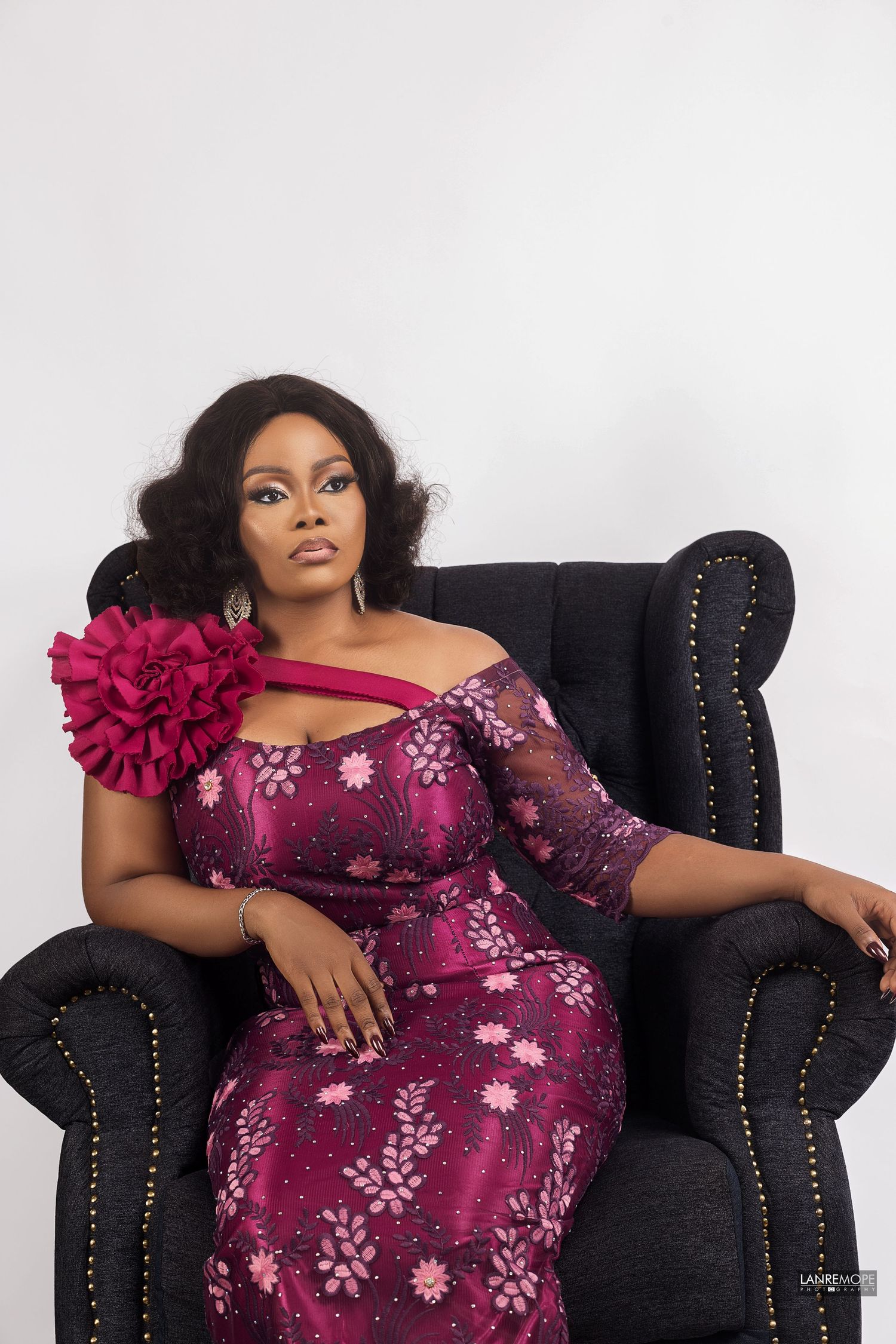 Woman in burgundy floral off-shoulder gown with large rose accent poses elegantly in black velvet armchair.