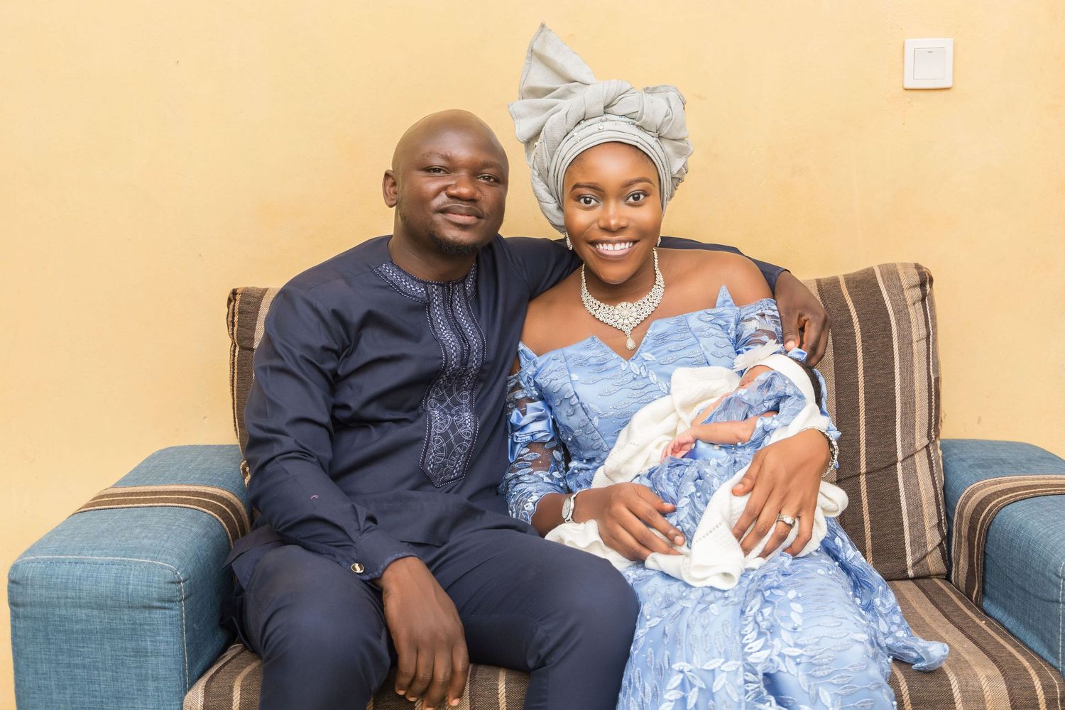 Nigerian couple in traditional blue attire sitting on sofa holding their newborn baby.