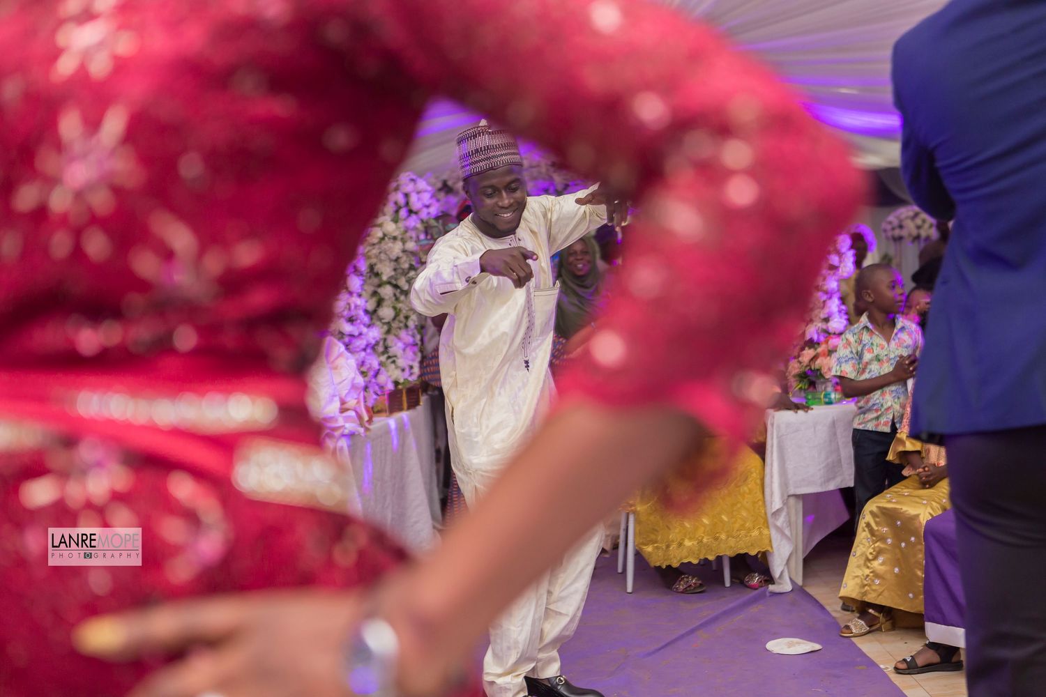 Man in white traditional attire dancing joyfully at a vibrant African wedding celebration with colorful decor.