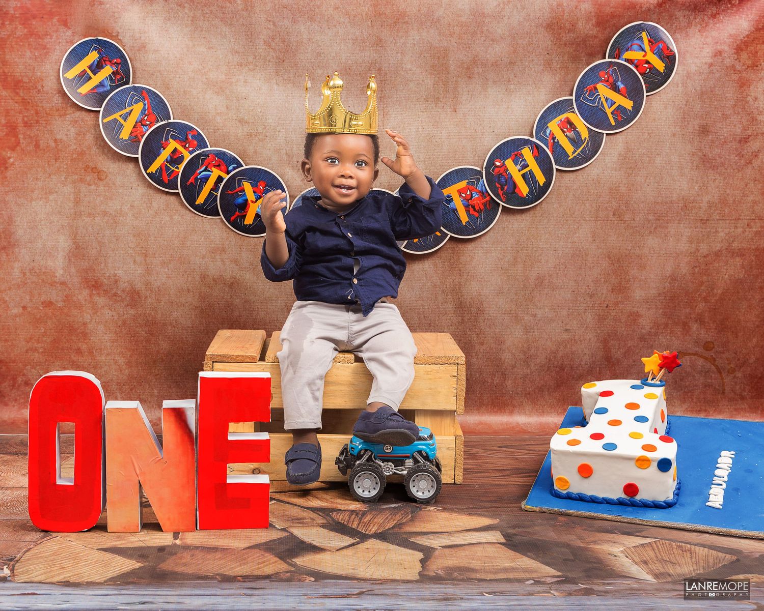 Toddler boy wearing a gold crown sitting on a crate with ONE letters and a birthday cake.