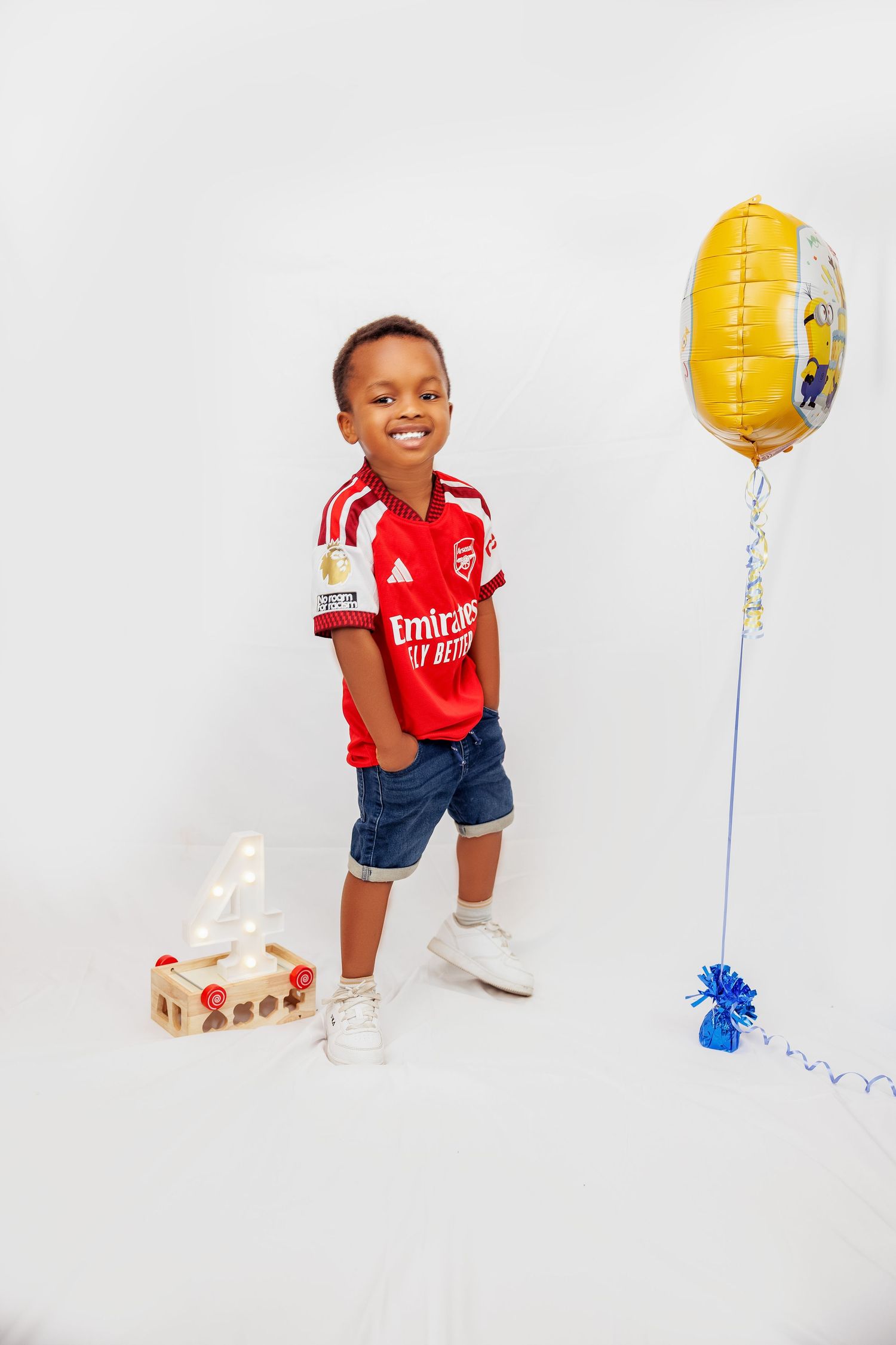 Happy boy in Arsenal jersey posing with gold birthday balloon and number 4 block on white background.