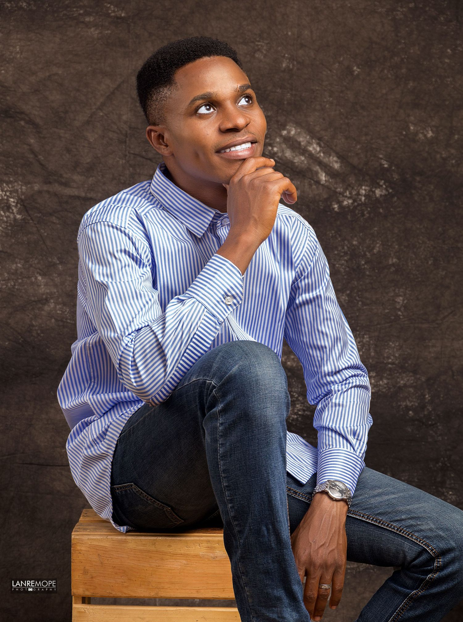 Young man in blue striped shirt and jeans posing thoughtfully in a professional studio portrait.