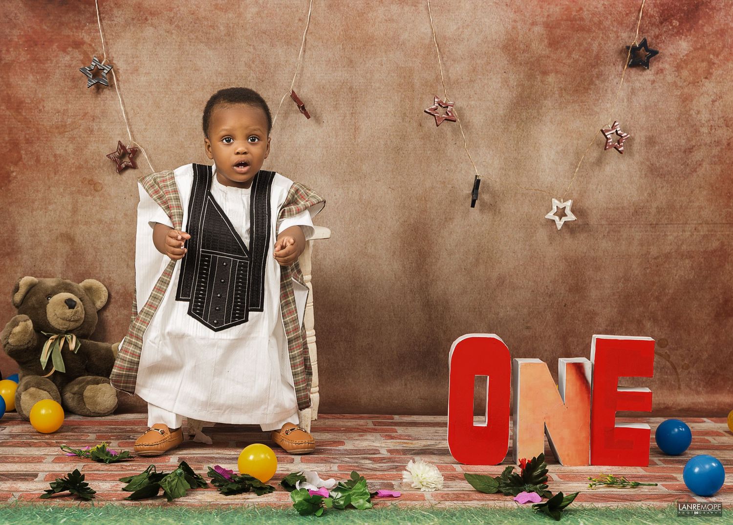 Toddler boy in formal vest outfit standing next to red ONE letters in a decorated studio.