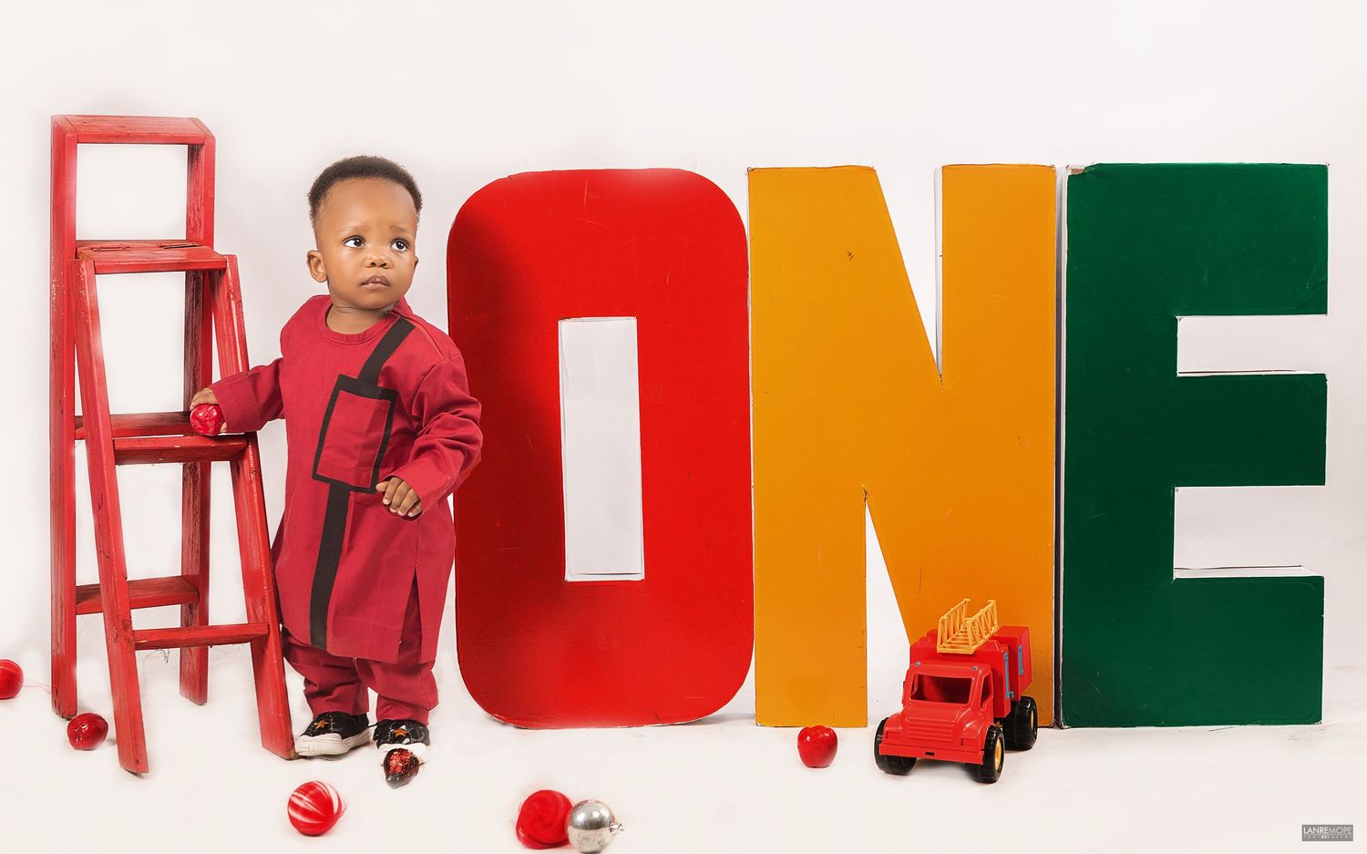 Toddler boy in red outfit standing beside large colorful ONE letters with toy truck for birthday.
