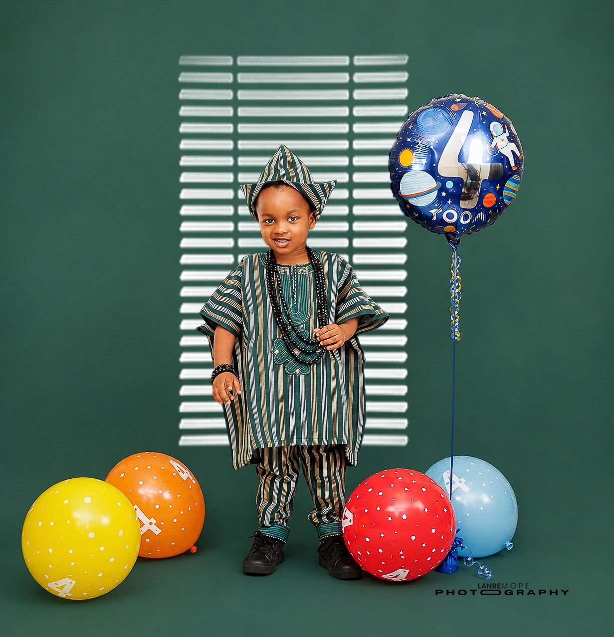 Young boy in traditional African agbada outfit posing with colorful balloons for 4th birthday photos.