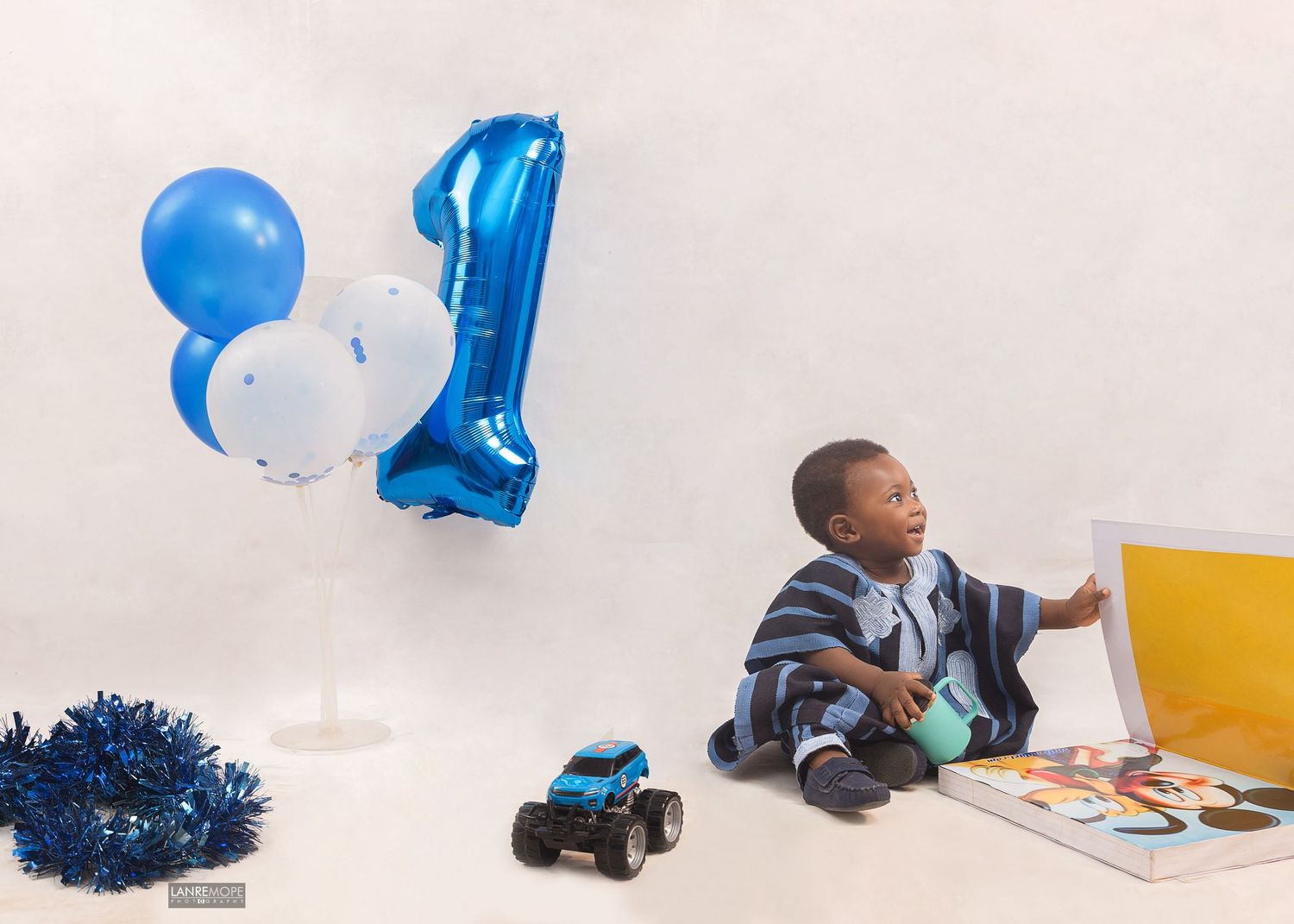 Young boy sitting beside a large blue number 1 balloon with toys on his first birthday.