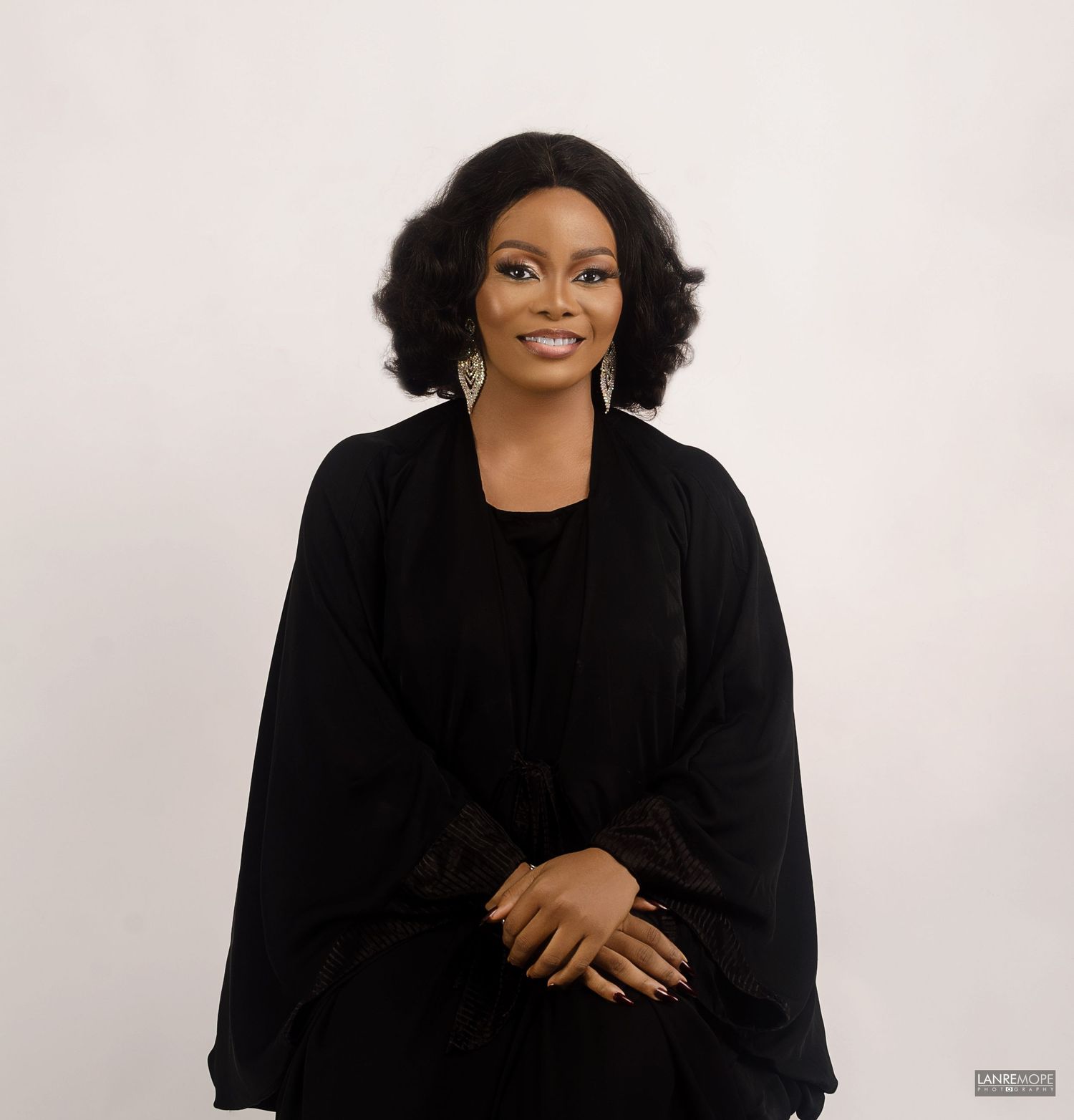 Woman in elegant black outfit and statement earrings smiling confidently against a white background.