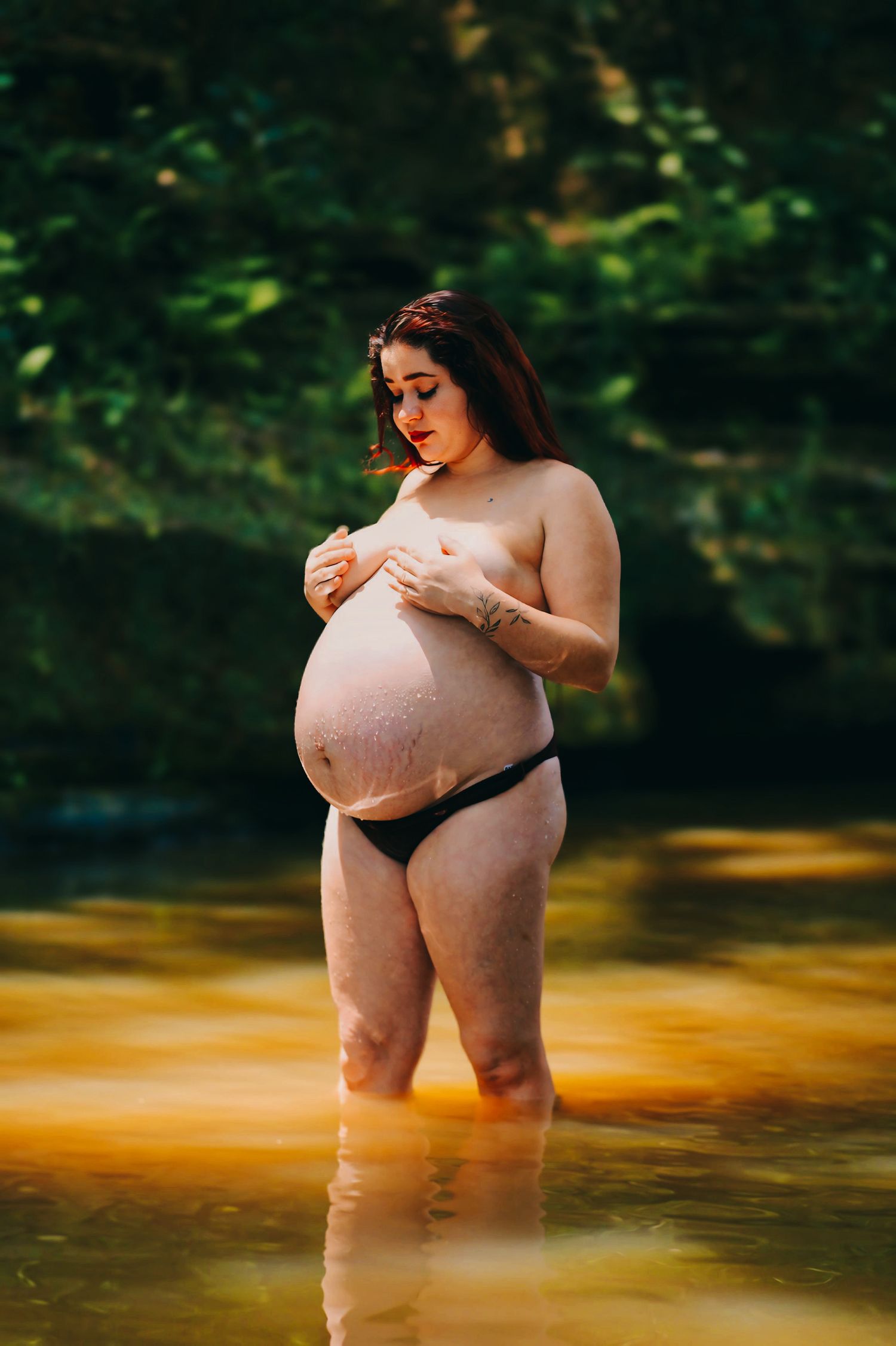 Gestante em ensaio fotográfico artístico dentro de um rio, com as mãos sobre o peito.