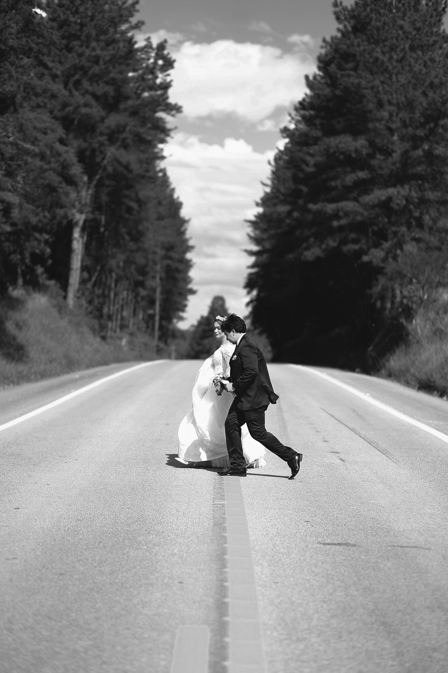 Casal de noivos atravessando uma estrada de mãos dadas.