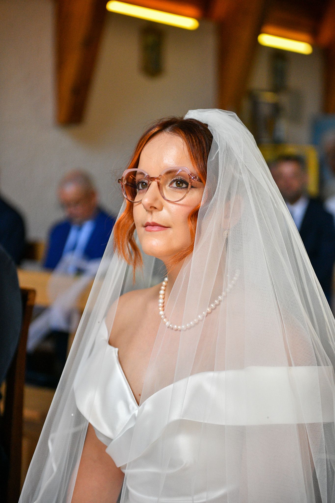 Red-haired bride wearing glasses and pearl necklace under a white wedding veil.