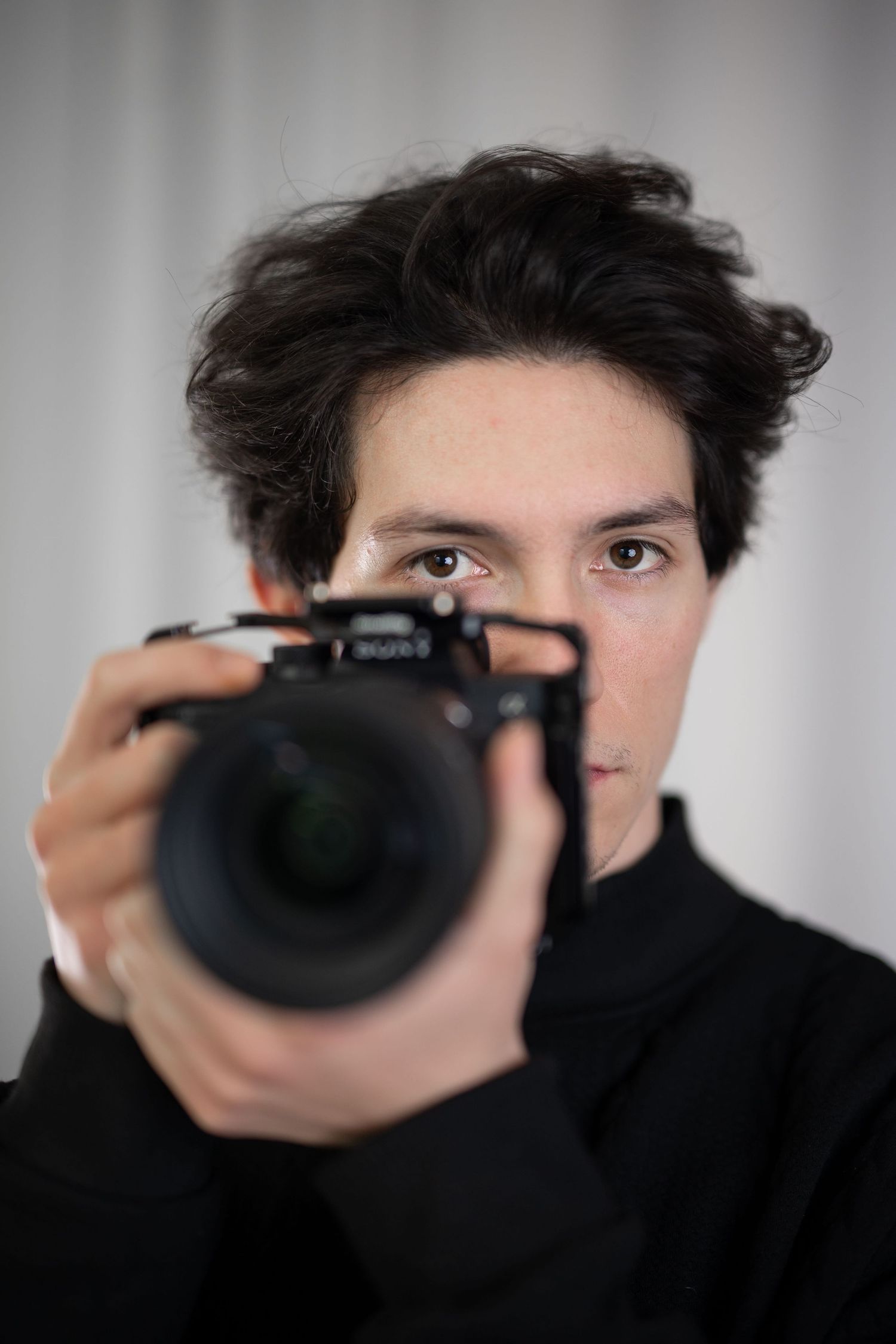 Young photographer holding a professional DSLR camera up to their face against a gray background.
