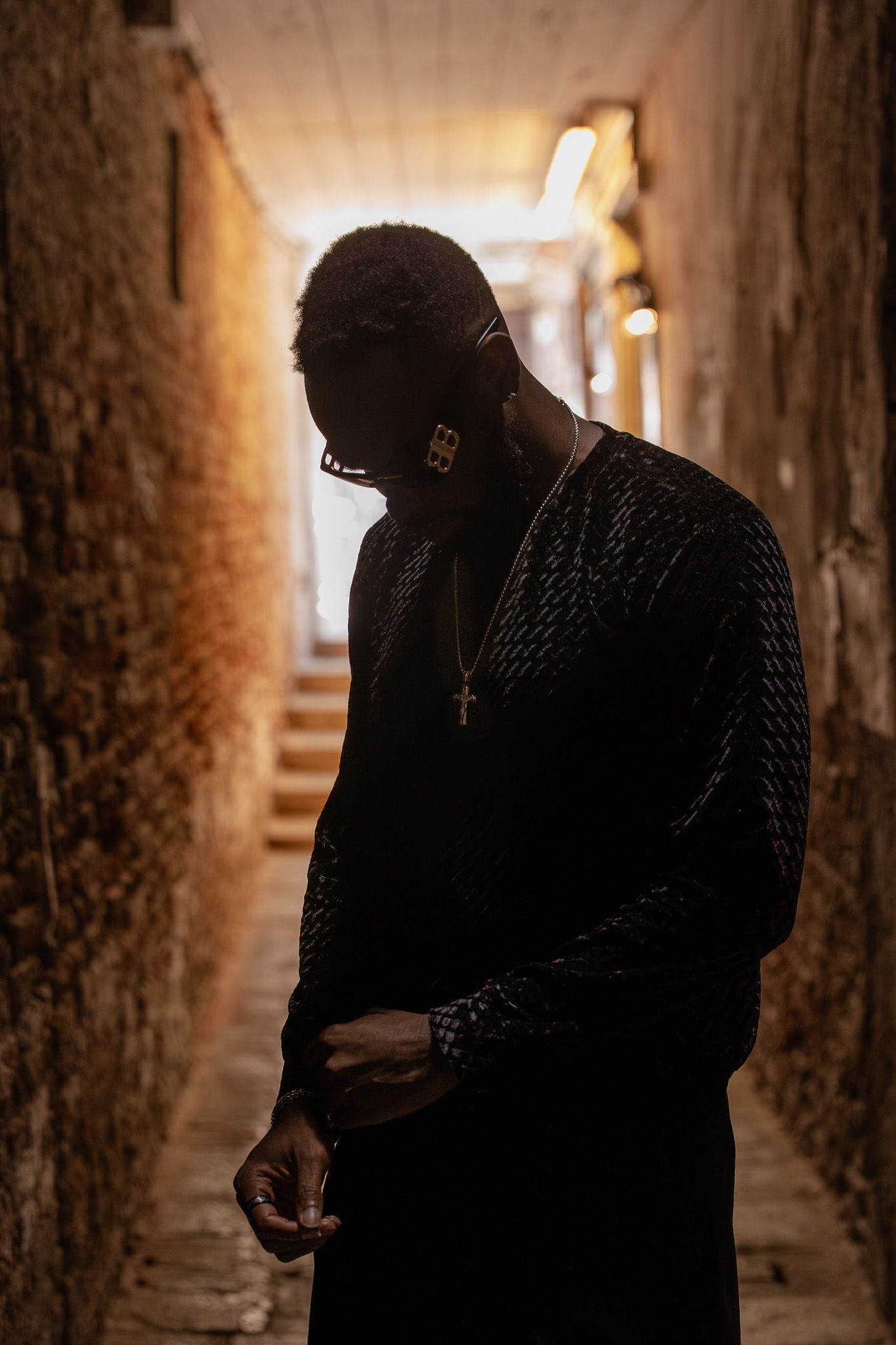 Man in black textured outfit with gold cross necklace stands in dimly lit brick corridor with light at end.