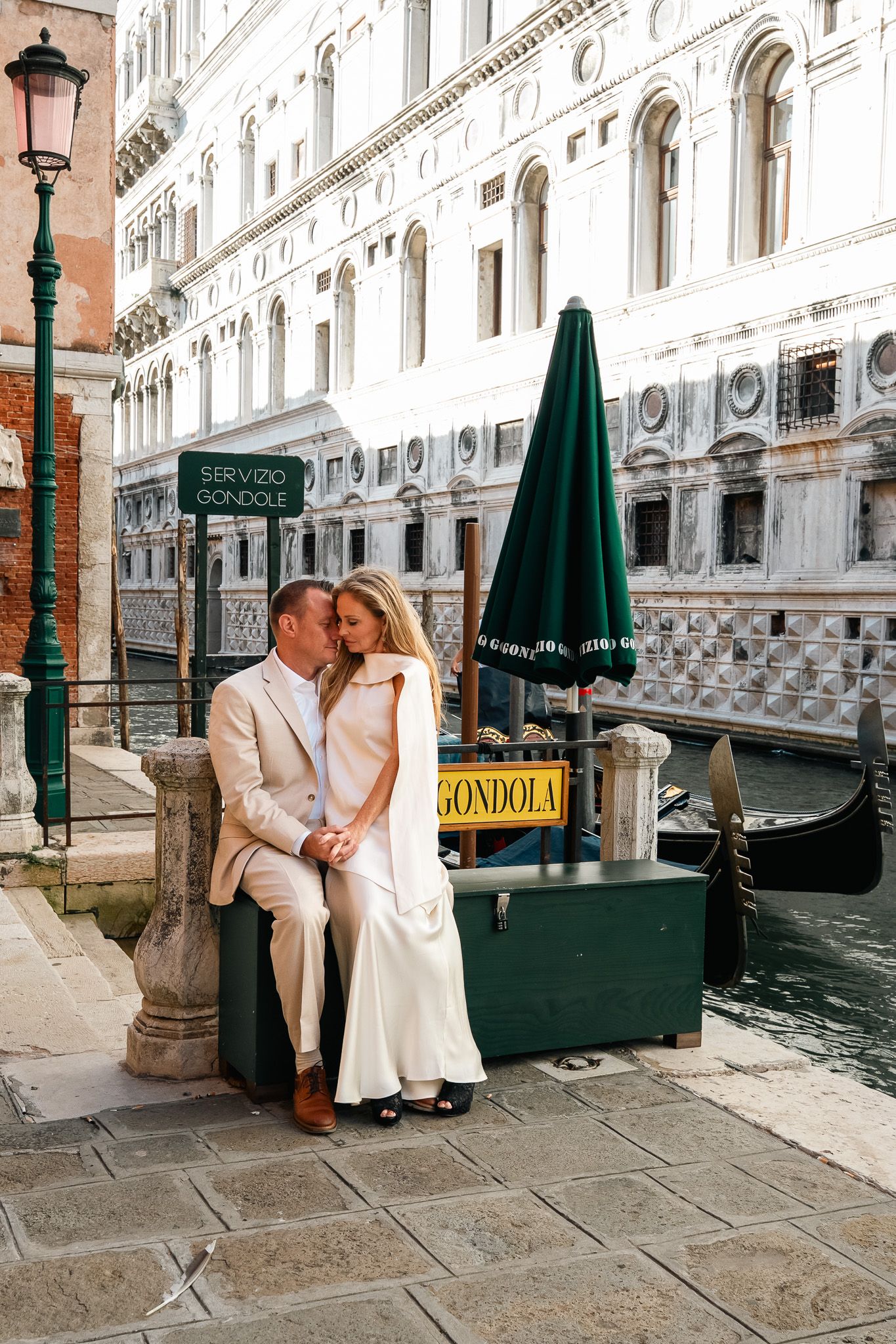 Couple sharing a romantic kiss by a gondola station on a Venice canal, dressed in elegant white attire.