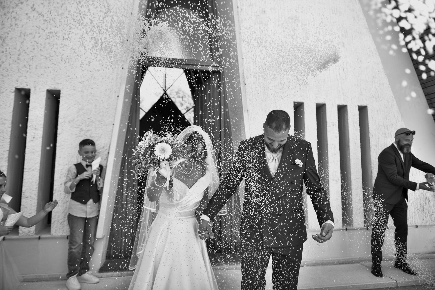 Black and white photo of bride and groom showered with confetti outside church.