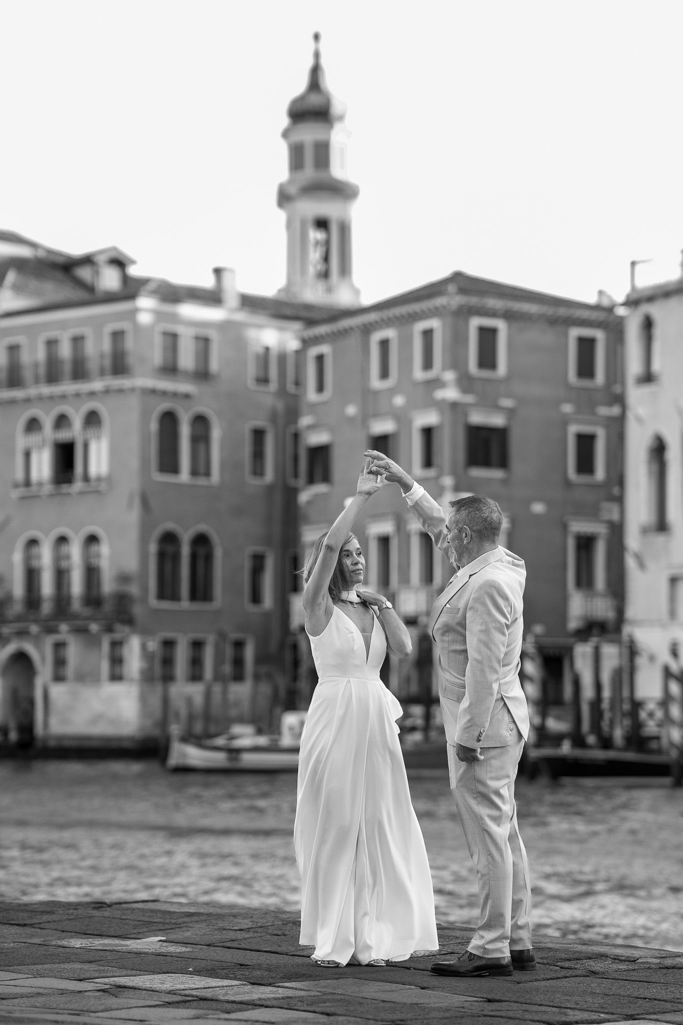 Couple dancing on Venice waterfront in black and white, woman in white dress, man in suit, historic buildings behind.