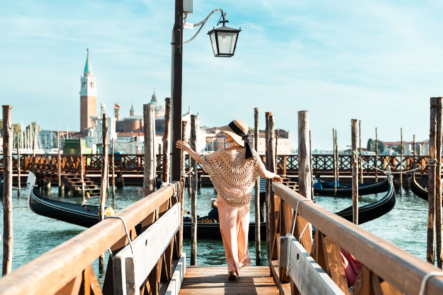 Woman in hat walking along Venice gondola dock with San Giorgio Maggiore backdrop.