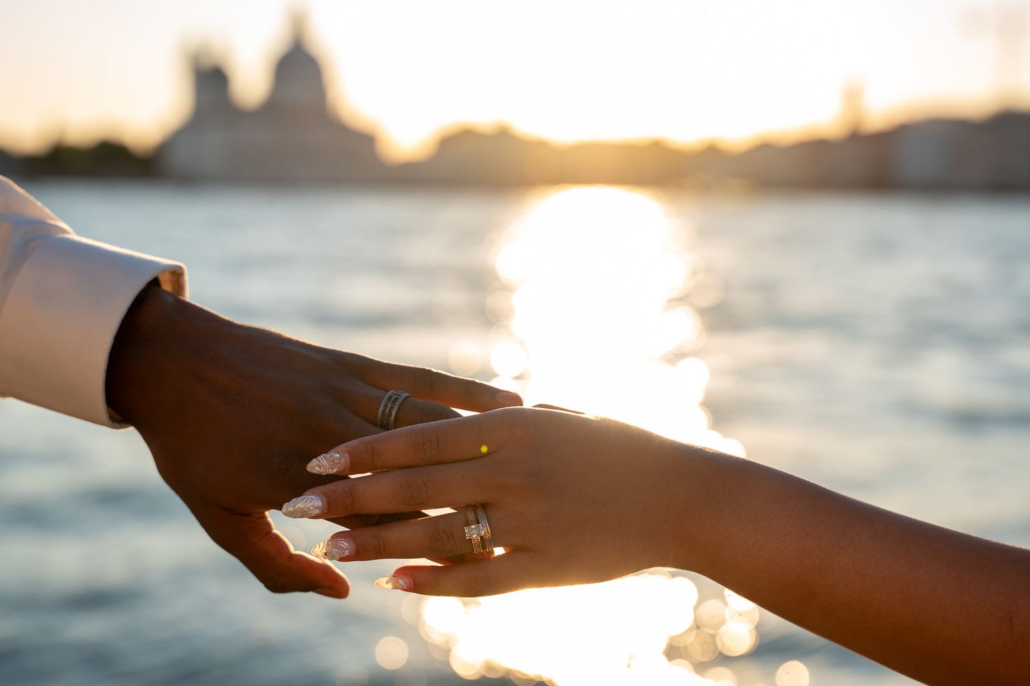 Two hands touching at sunset over water, one wearing a diamond engagement ring, with a romantic cityscape silhouette in background.