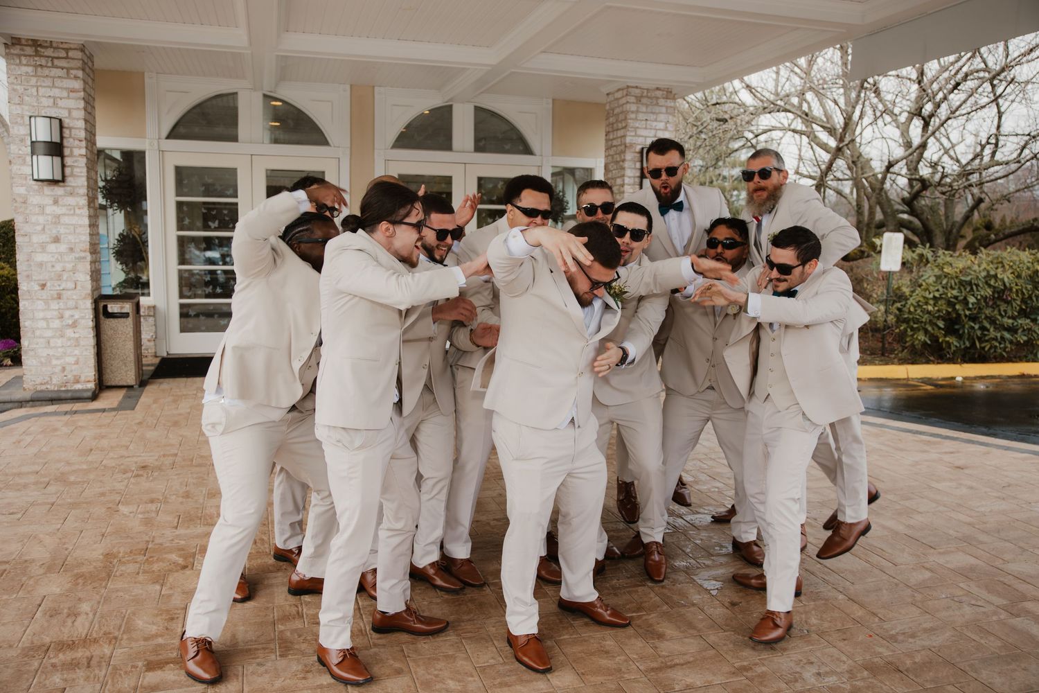 Groomsmen in white suits and brown shoes pose playfully outside a venue for a wedding photo.