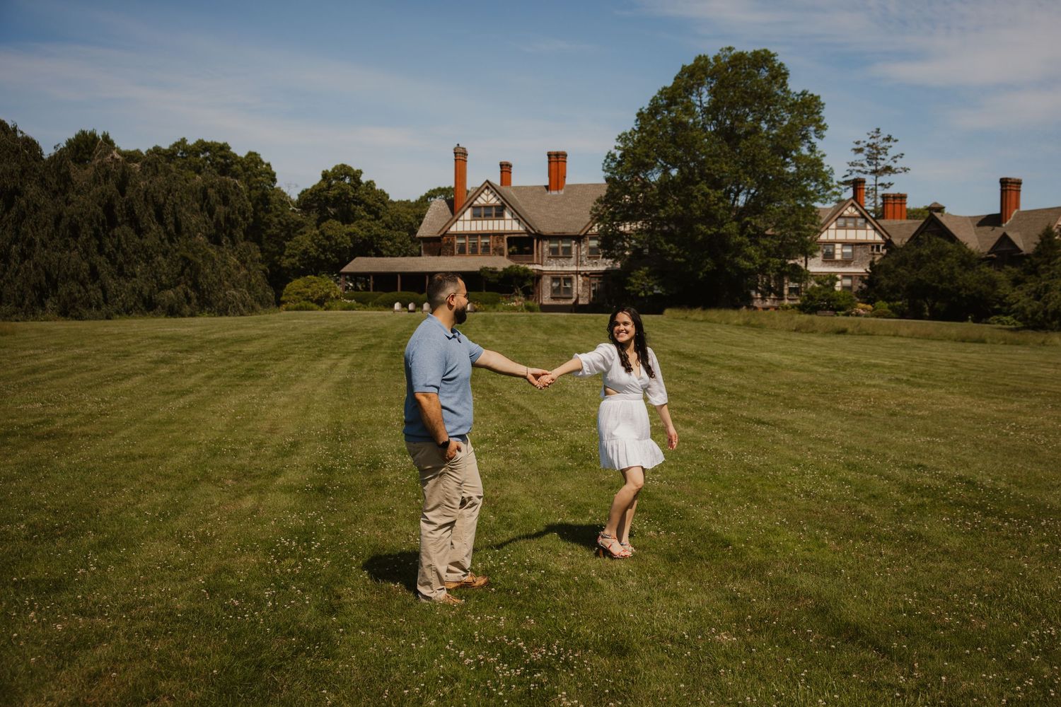 An engaged couple holds hands and dances playfully on a vast green lawn in front of a historic estate.