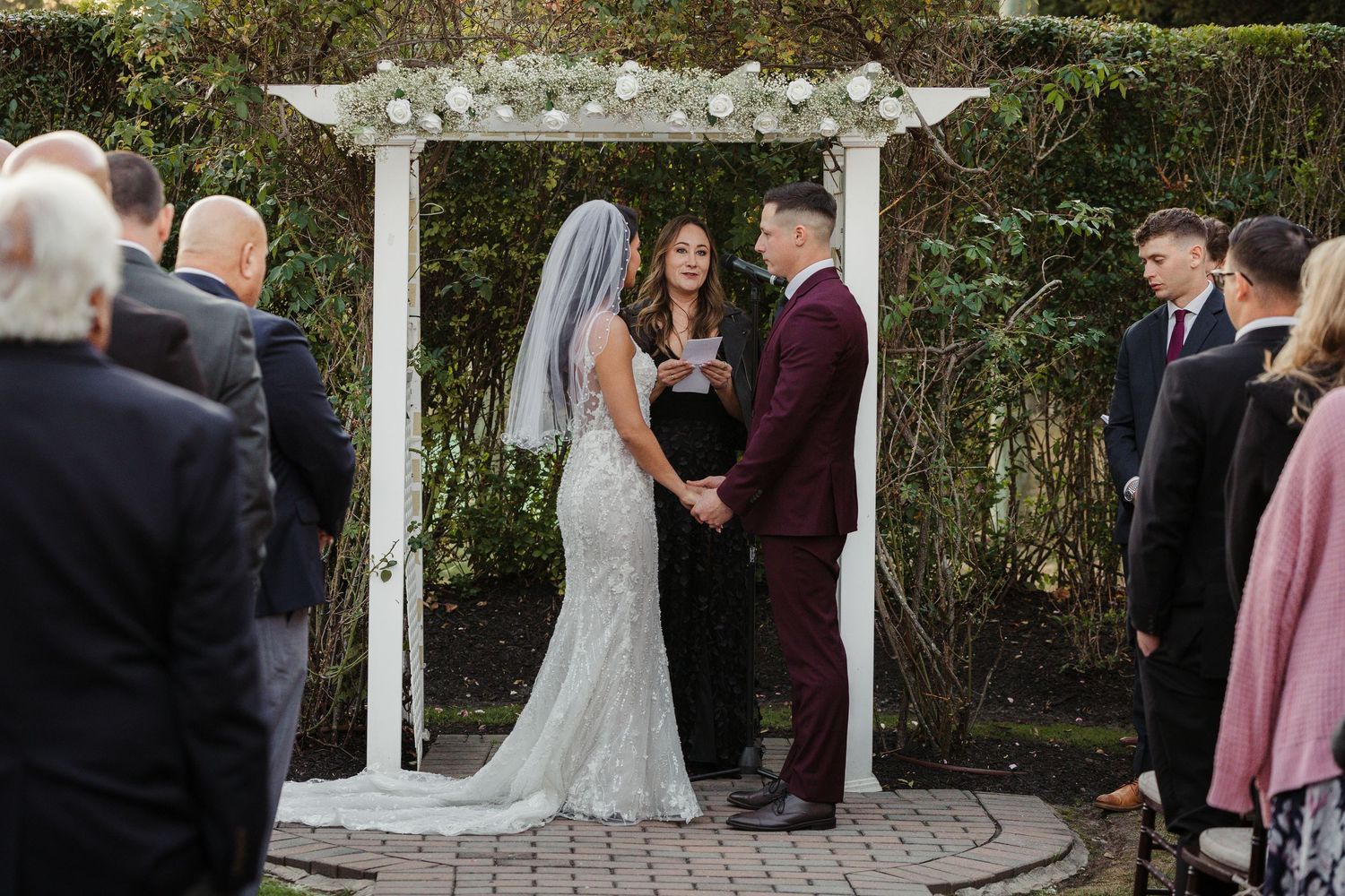 Bride and groom exchange vows under a white floral arbor during an outdoor wedding ceremony with guests watching.