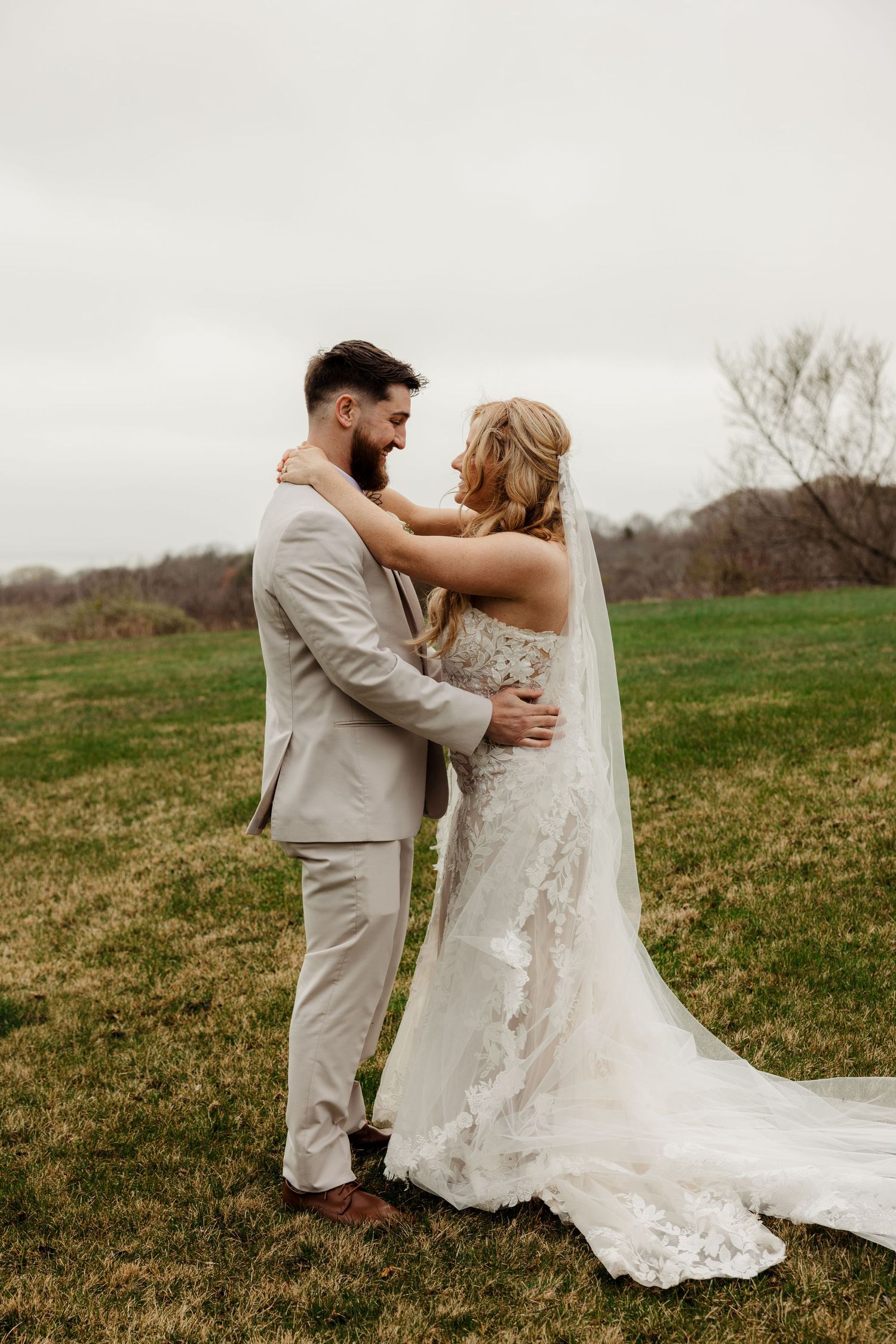 Bride and groom embrace in an open field wearing lace wedding gown and tan suit on a cloudy day.