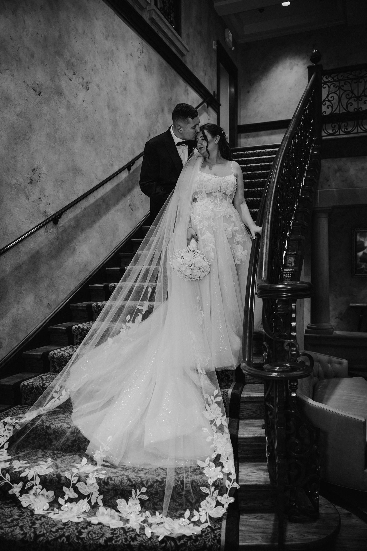 Black and white photo of bride and groom on an elegant staircase with ornate iron railing.