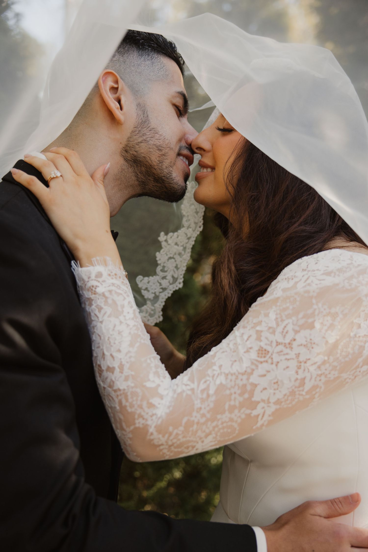Bride and groom share an intimate nose-to-nose moment under flowing white veil outdoors.
