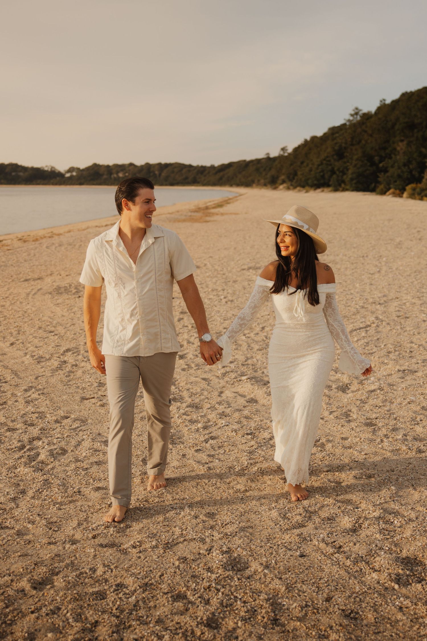 Couple in white outfits holding hands and walking barefoot on a sandy beach at golden hour.