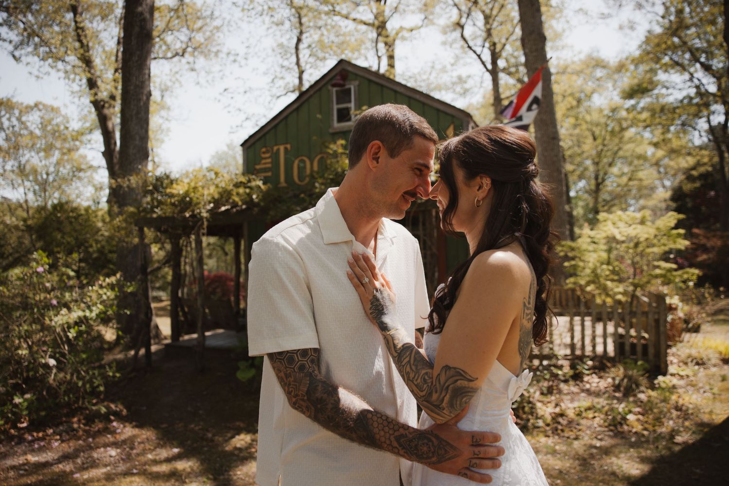 Tattooed couple embrace and lean foreheads together near a rustic green cabin in the woods.