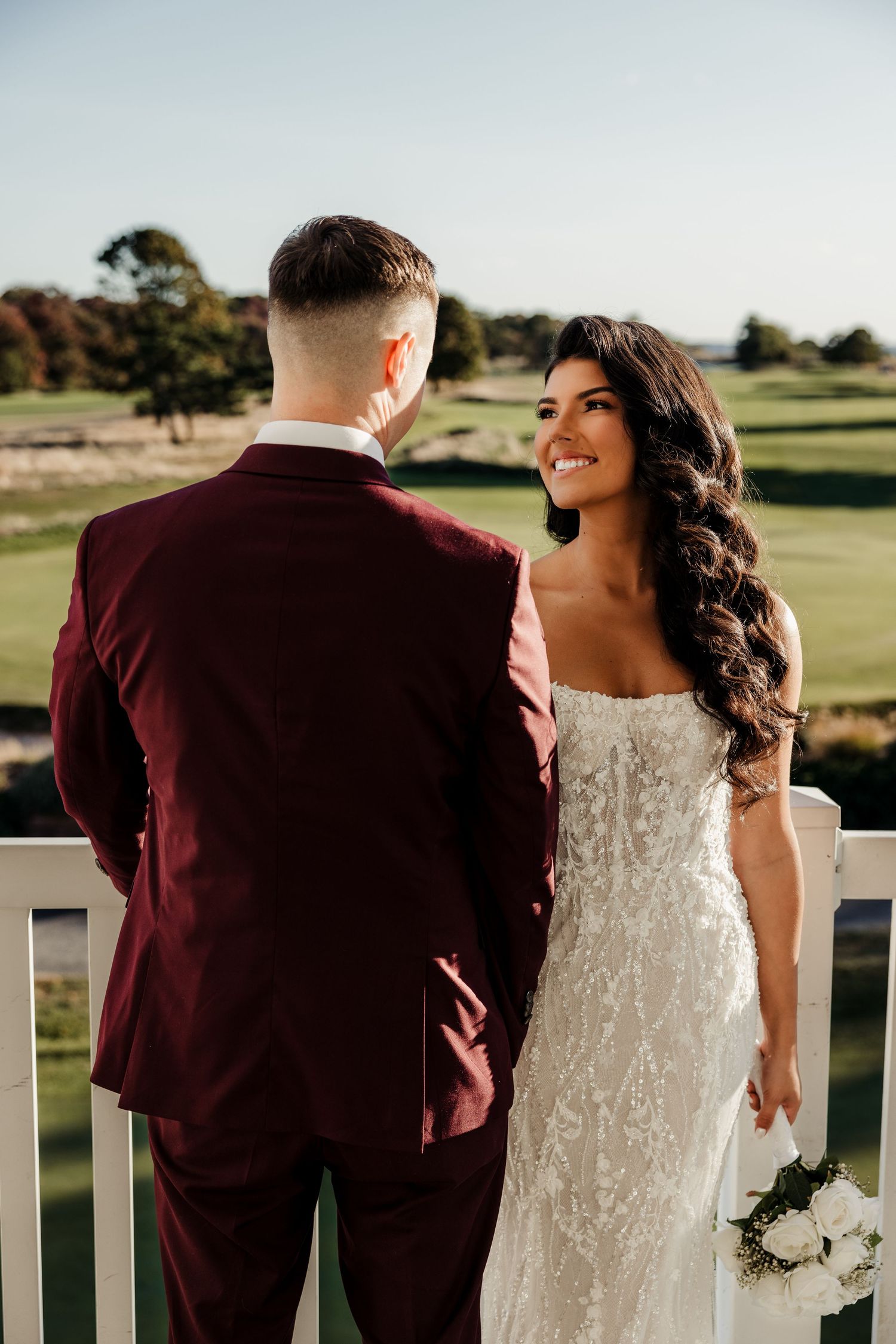 Smiling bride in lace gown with groom in burgundy suit on a golf course balcony.